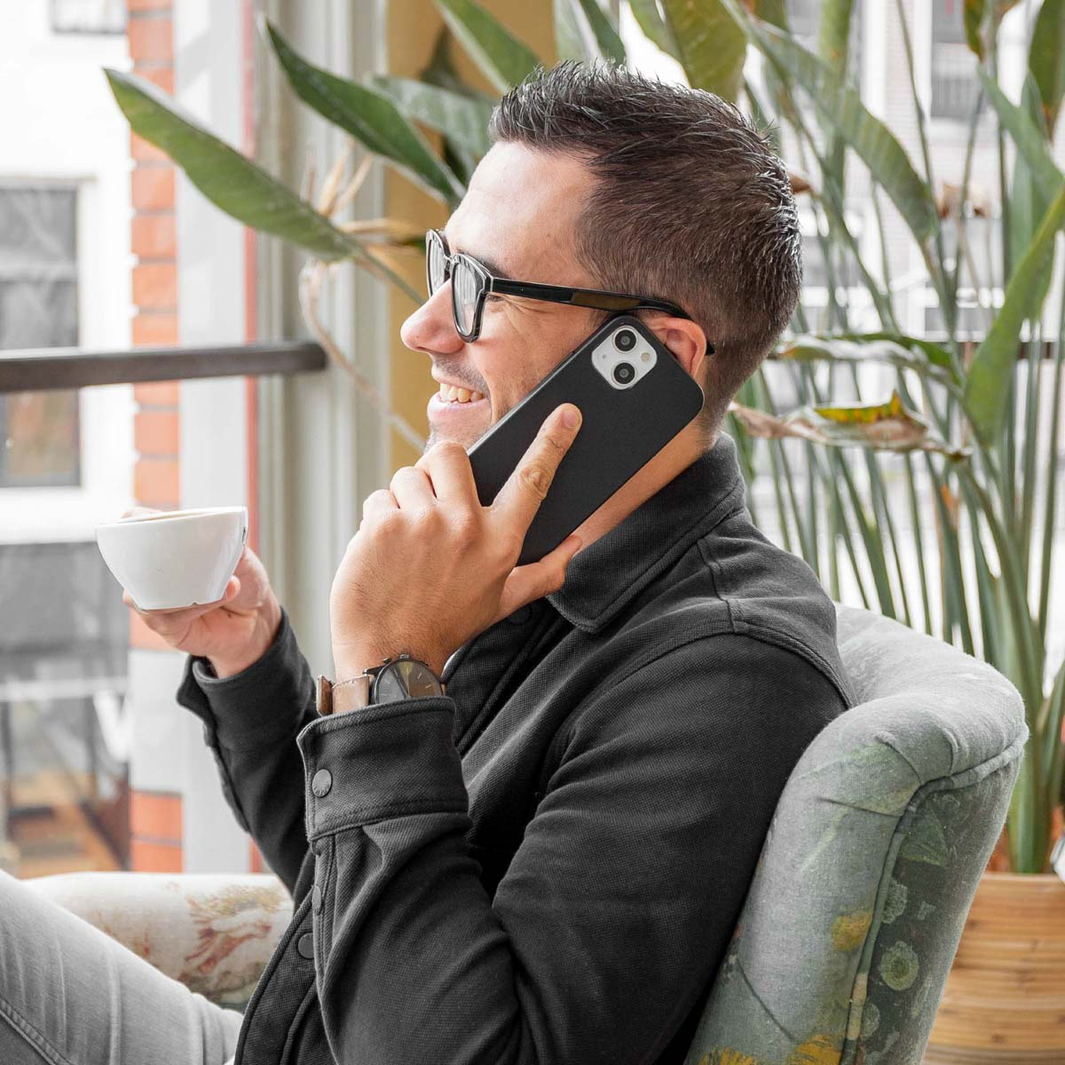 Een man gebruikt een zwarte telefoon, lachend, met een kopje koffie, binnen met planten.