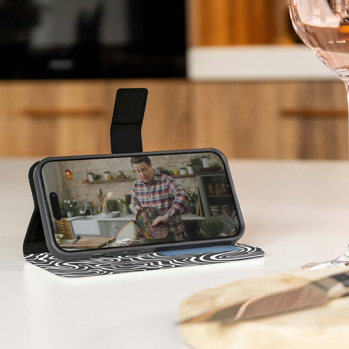 Telefoon in een hoesje met een video van een persoon die kookt, op een tafel met een wijnglas op de achtergrond.