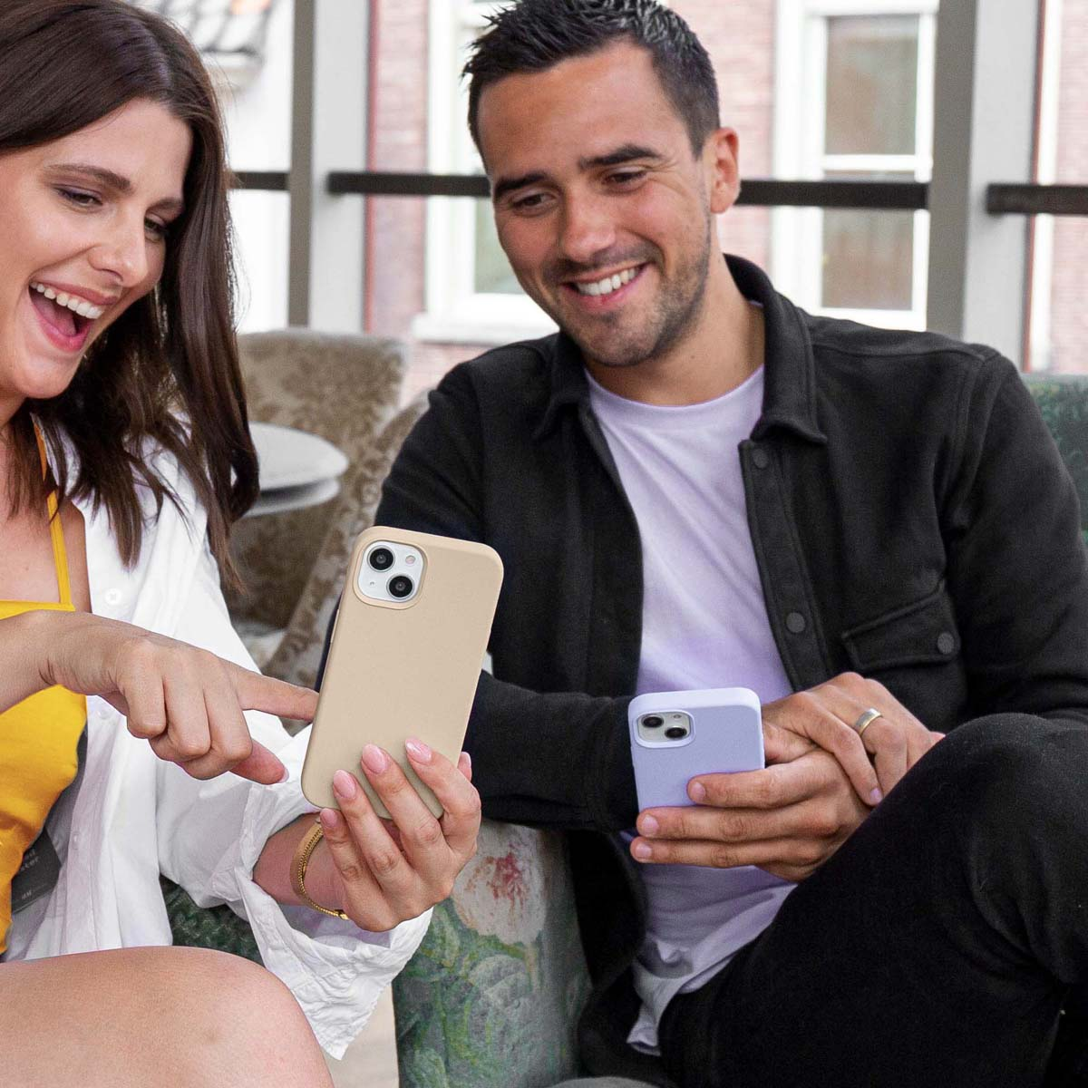 Twee mensen kijken naar telefoons, één met een beige hoesje, de ander een paars. Beide lachen.