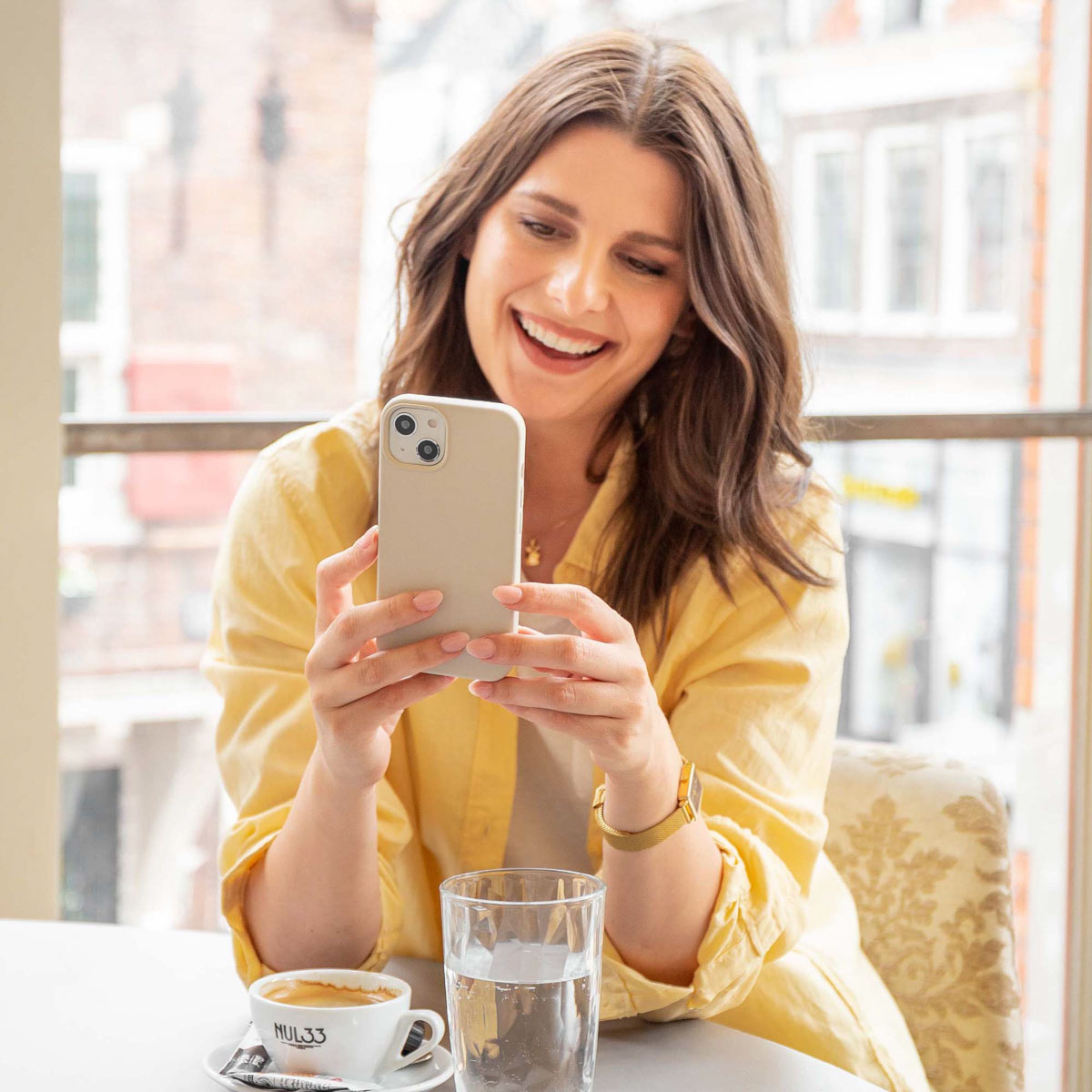 Vrouw lacht terwijl ze naar telefoon kijkt. Koffie en water op tafel.