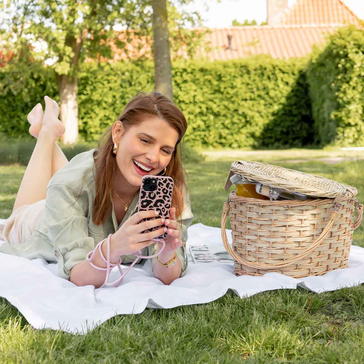 Eine lächelnde Frau hält ihr Telefon mit einer Leopardenmuster-Hülle. Ein Picknickkorb und eine Decke sind im Bild.