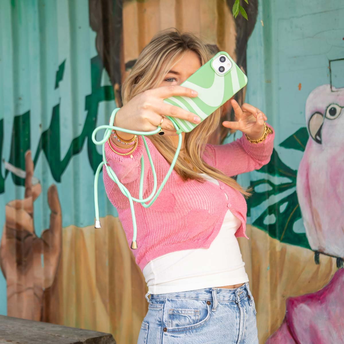 Een blonde vrouw maakt een selfie met een lichtgroene telefoon voor een kleurrijke muur.