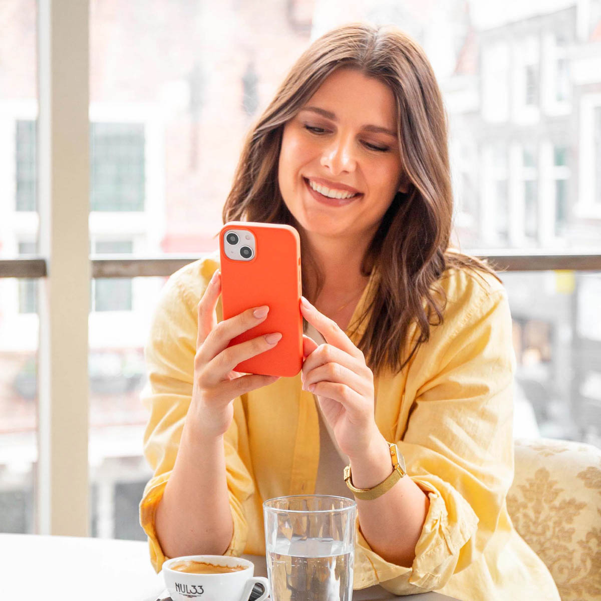Vrouw met bruin haar, houdt een oranje telefoonhoes vast, glimlacht. Achtergrond: raam, koffiekopje, waterglas.