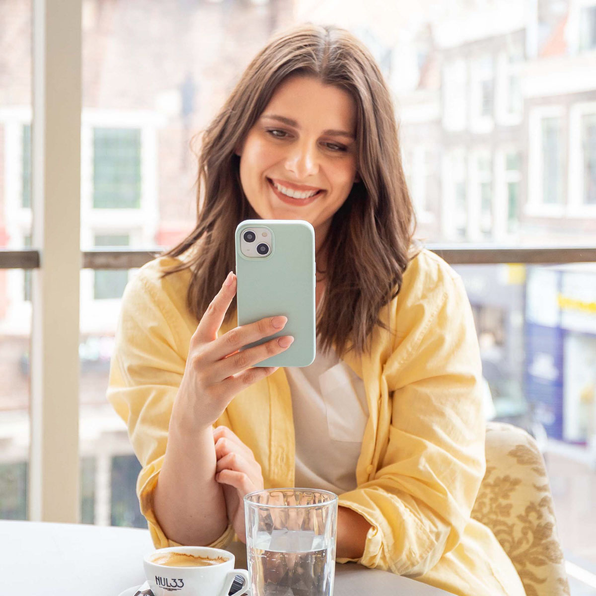 Vrouw lacht, houdt een groene telefoon vast, bij een raam met een kopje koffie en water.