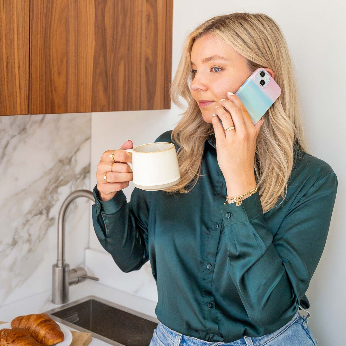 Vrouw aan de telefoon, houdt een mok vast. Achtergrond: kraan, marmeren muur en croissants op een bord.