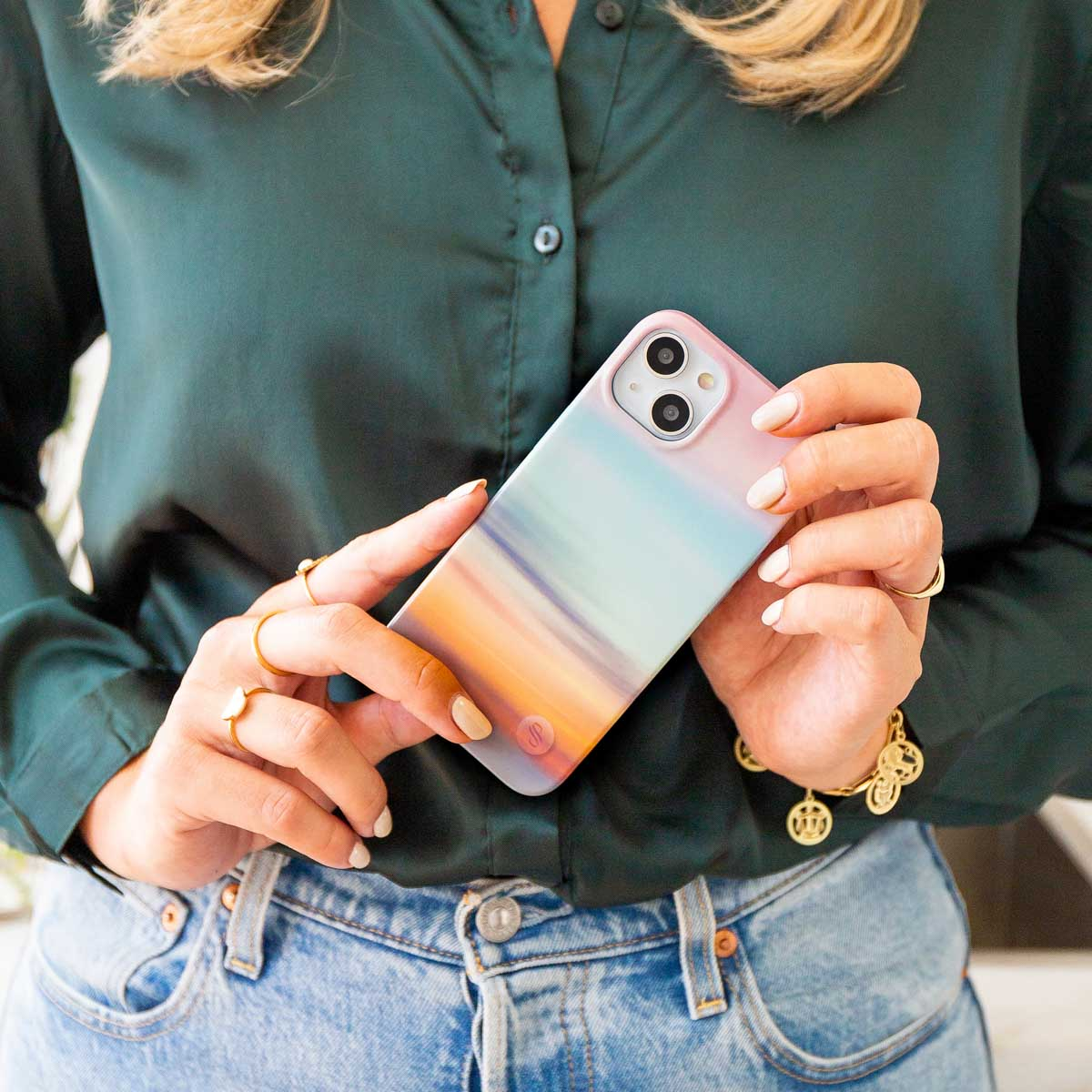 Vrouw met telefoon met kleurrijke hoes, met ringen aan haar vingers, jeans en groen shirt.