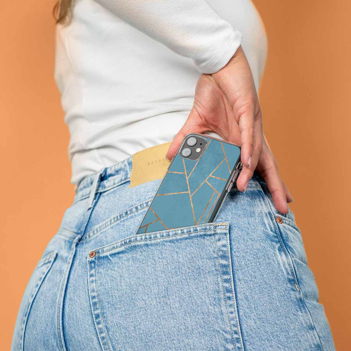 Een hand die een blauwgroene telefoonhoes in een jeanszak steekt. De hoes heeft een gouden design.