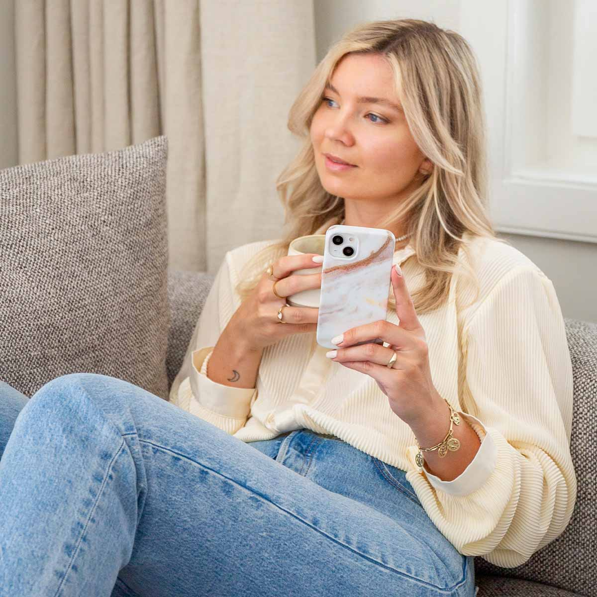 Femme sur un canapé tenant un téléphone avec une coque en marbre et une tasse. Elle regarde ailleurs.