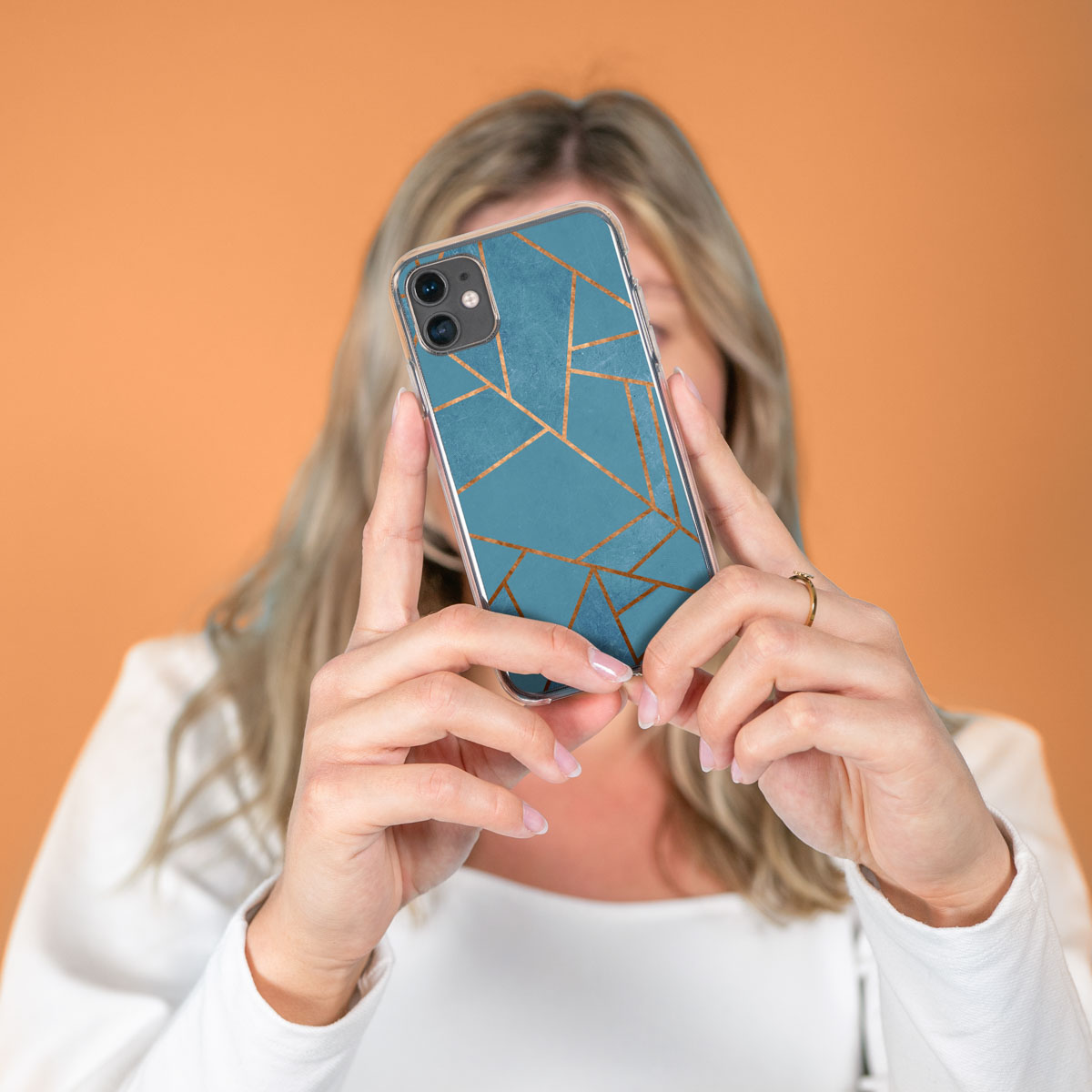 Een vrouw houdt een telefoon vast met een doorzichtige hoes, met een blauw en goud geometrisch ontwerp, met een oranje achtergrond.