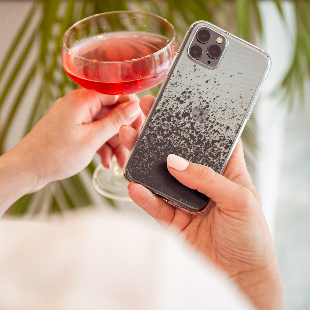 Une femme tient un verre avec une boisson rose et un téléphone. Le téléphone a une coque transparente avec des taches noires.