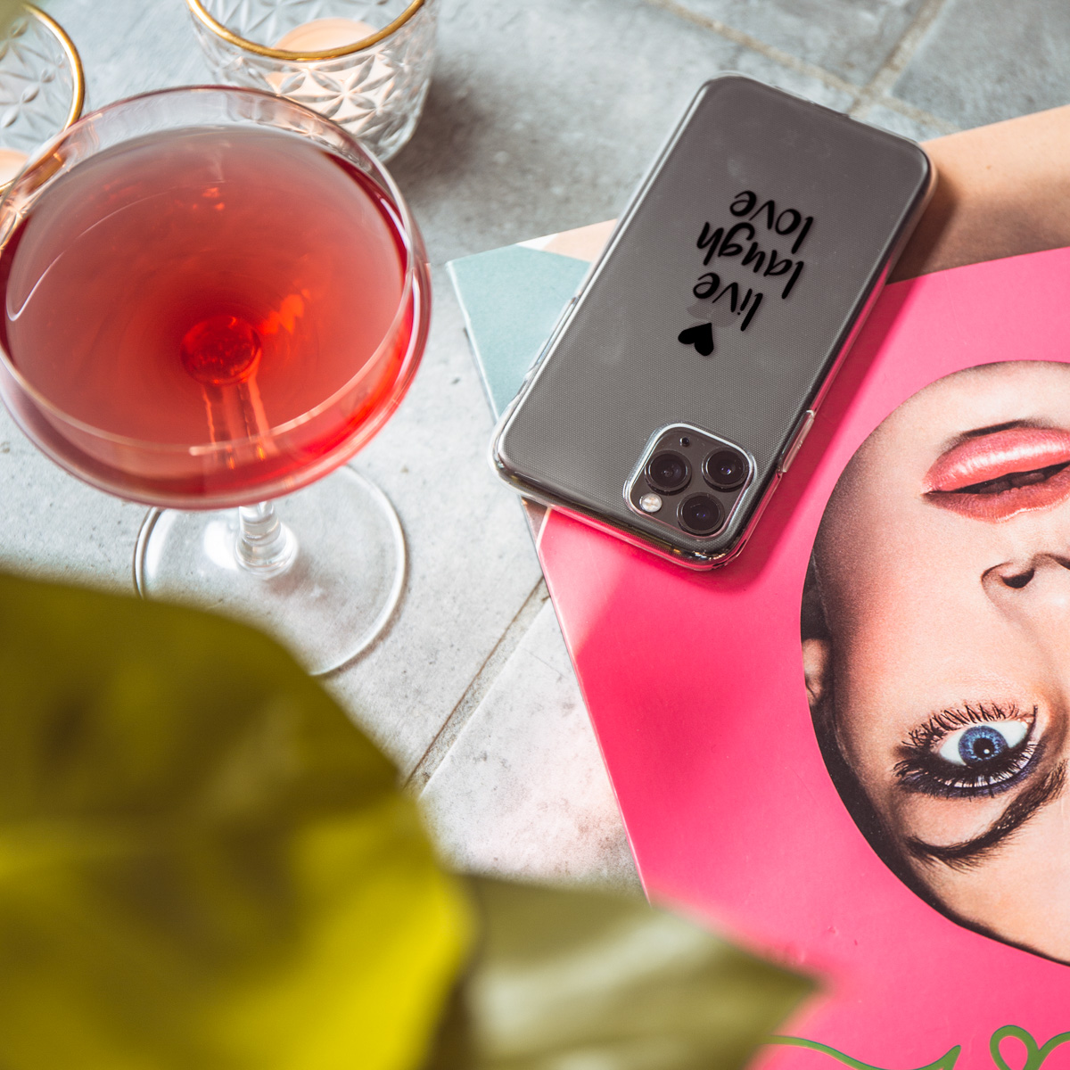 Telefoon in een doorzichtige hoes op een oppervlak met een drankje en bloemen. De telefoon zegt 'live laugh love'.