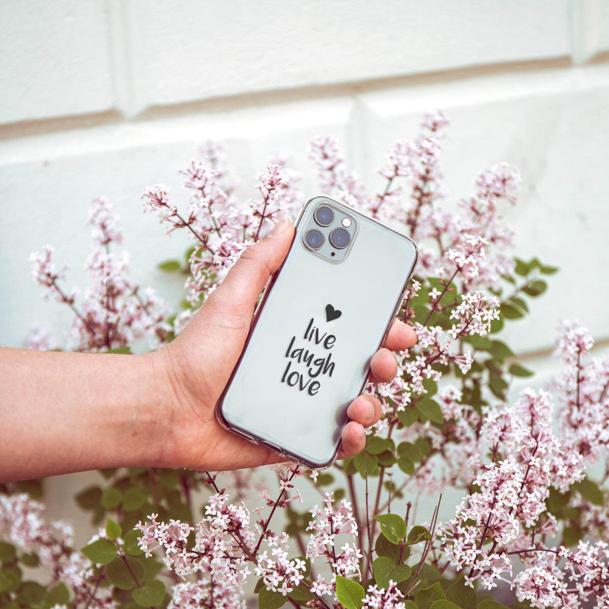 Hand die een telefoon vasthoudt met een doorzichtige hoes. De telefoon zegt 'live laugh love', bloemen op de achtergrond.