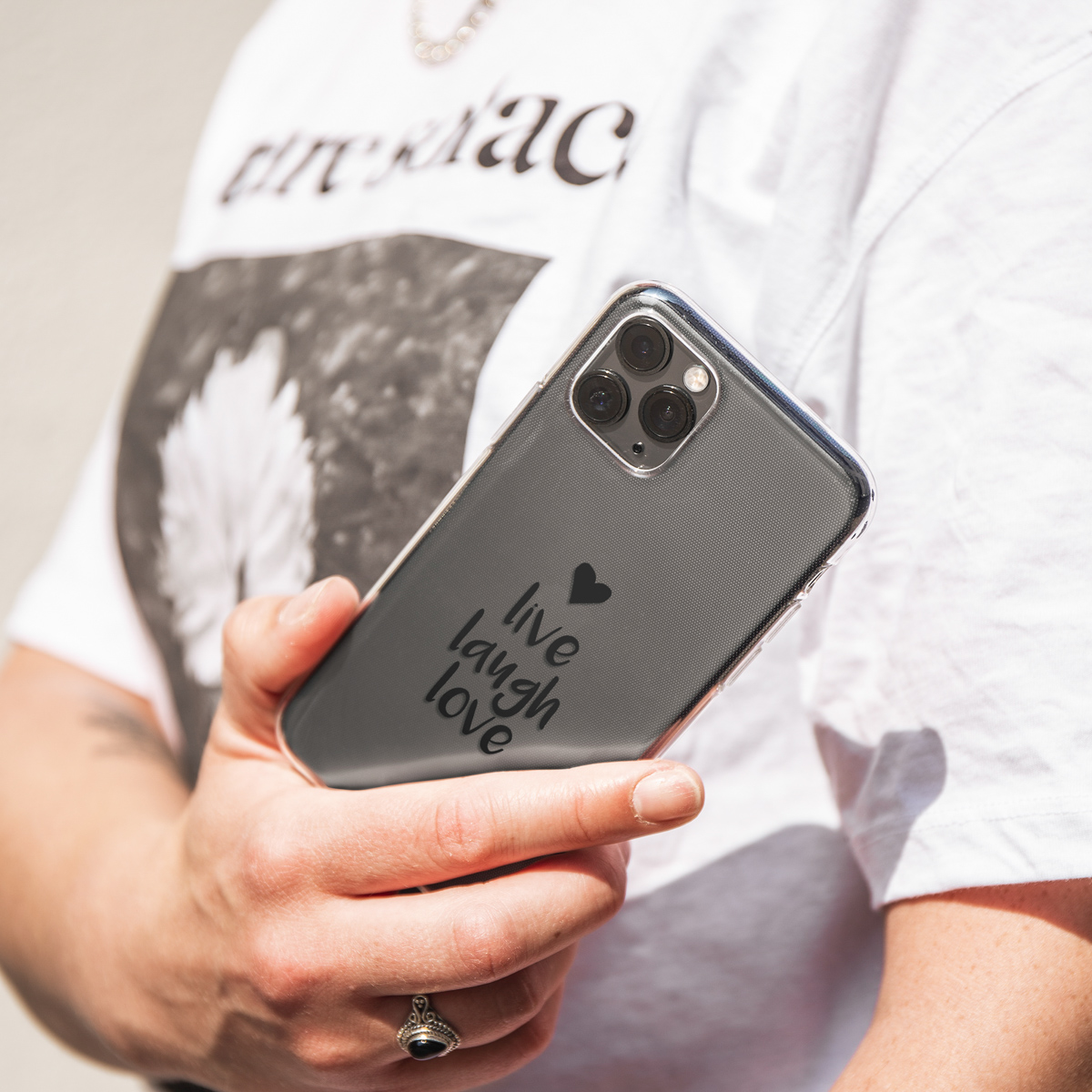 Persoon houdt telefoon met doorzichtige hoes vast. Telefoon zegt 'live laugh love'. Tekst is zwart op doorzichtige hoes.