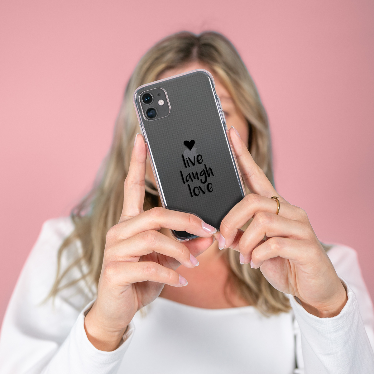 Vrouw houdt telefoon in doorzichtige hoes vast. Telefoon zegt 'live laugh love'. Roze achtergrond.