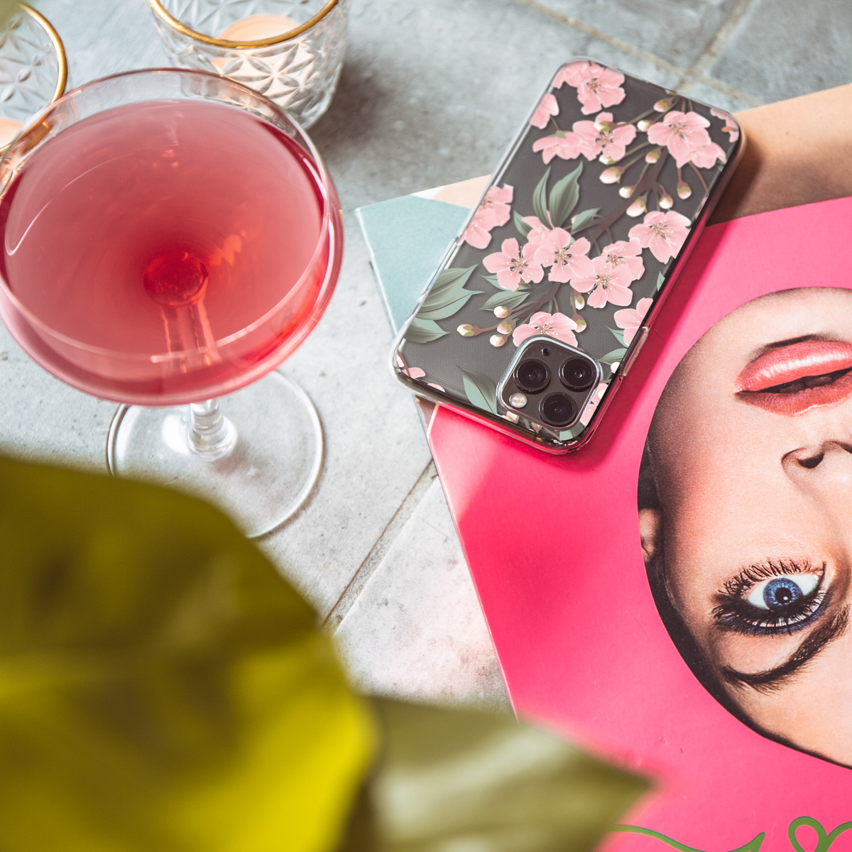 Telefoon in doorzichtige hoes met roze bloemen design, naast een drankje. Oppervlak is roze en grijs.