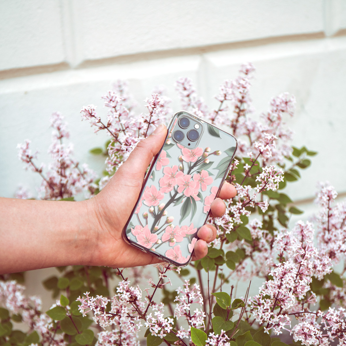 Hand die een doorzichtige telefoonhoes vasthoudt met roze bloemen. Achtergrond: witte muur en roze bloemen.