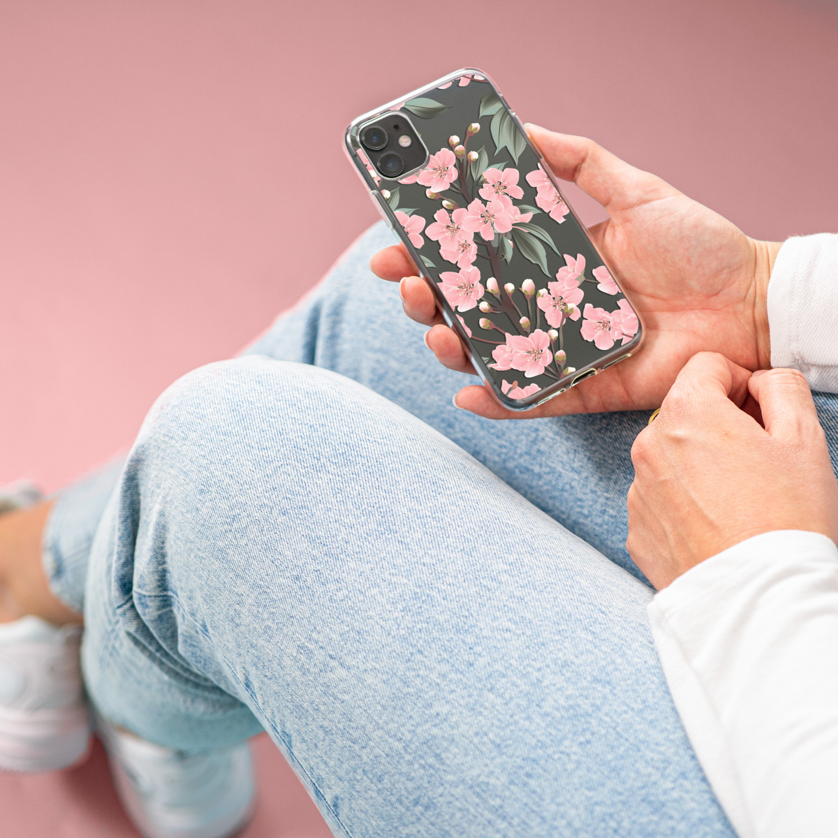 Persoon houdt telefoon met doorzichtige hoes, roze bloemen design vast. Zittend op denim, roze achtergrond.