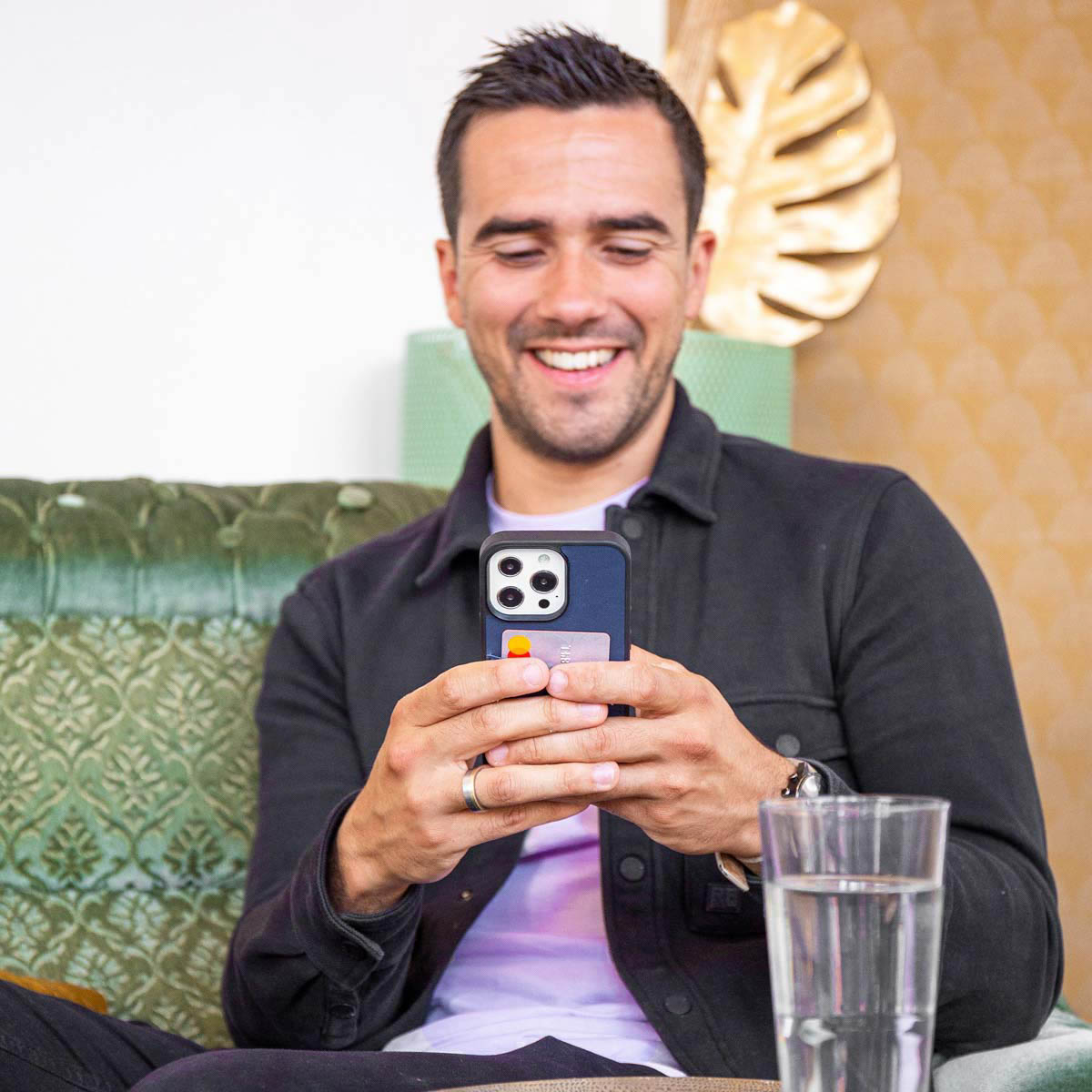 Homme tenant un téléphone avec une carte de crédit. Souriant. Verre d'eau sur la table. Canapé vert.