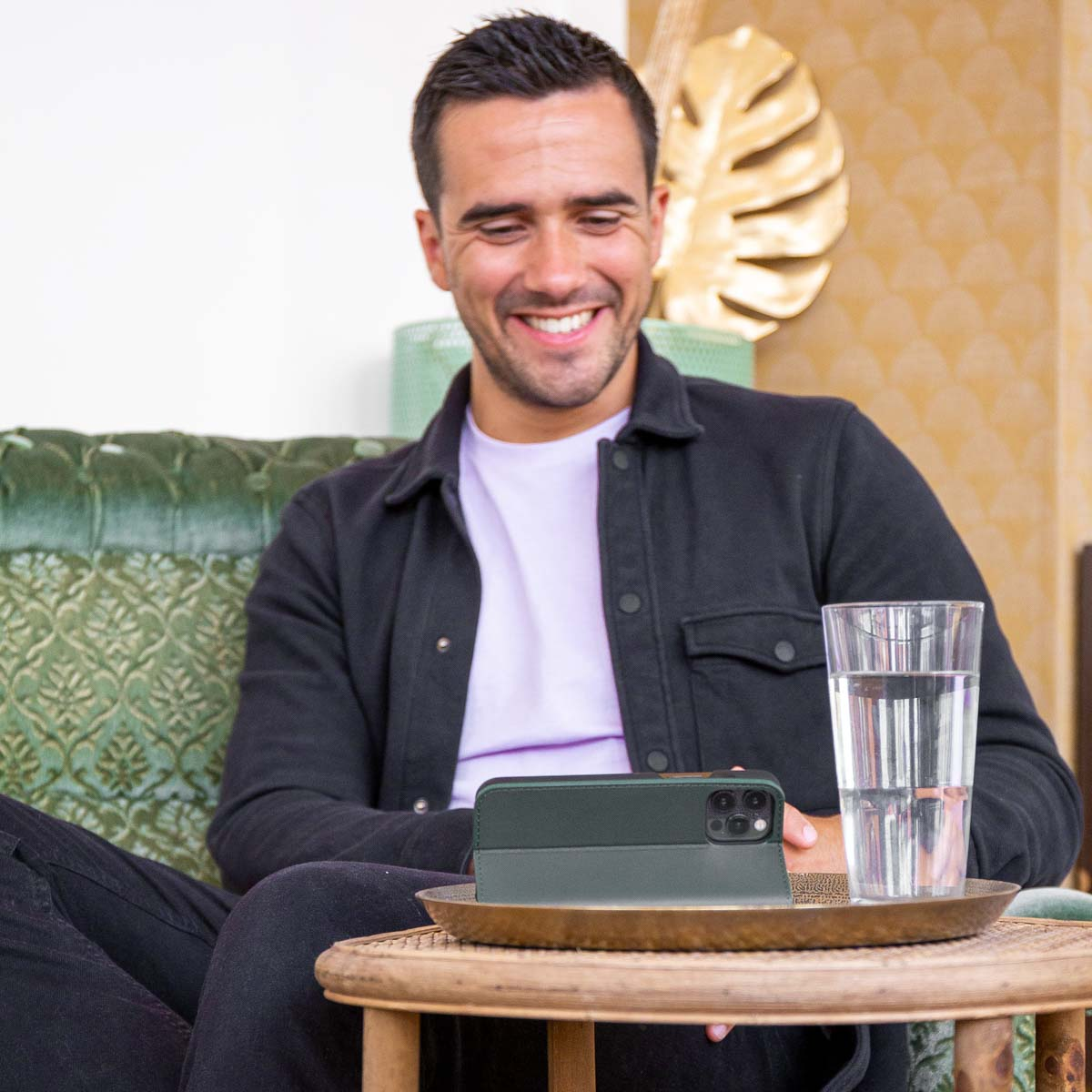 Homme souriant, assis sur un canapé vert, tenant un téléphone dans un étui. Arrière-plan flou.