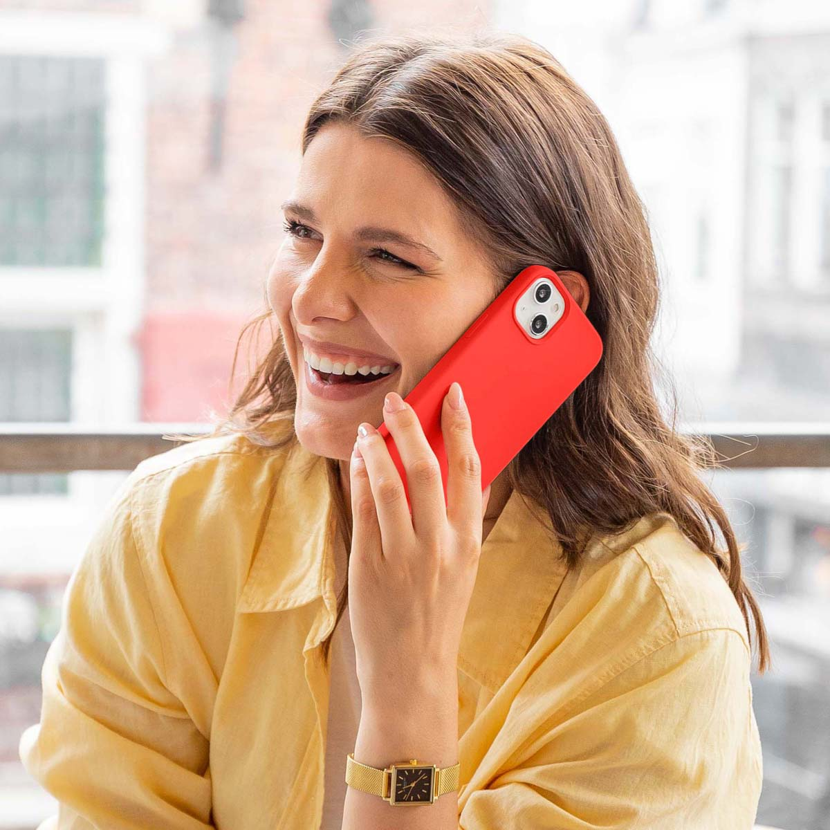 Une femme sourit en utilisant un téléphone rouge. Elle porte une chemise jaune et une montre dorée.