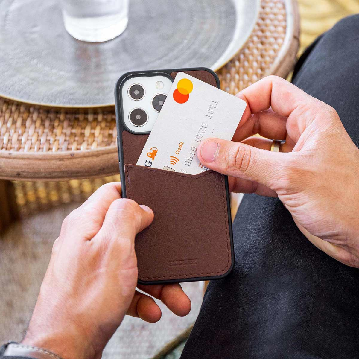 Persoon die een telefoon vasthoudt met een bruine hoes. Een creditcard wordt in een zakje gestoken.