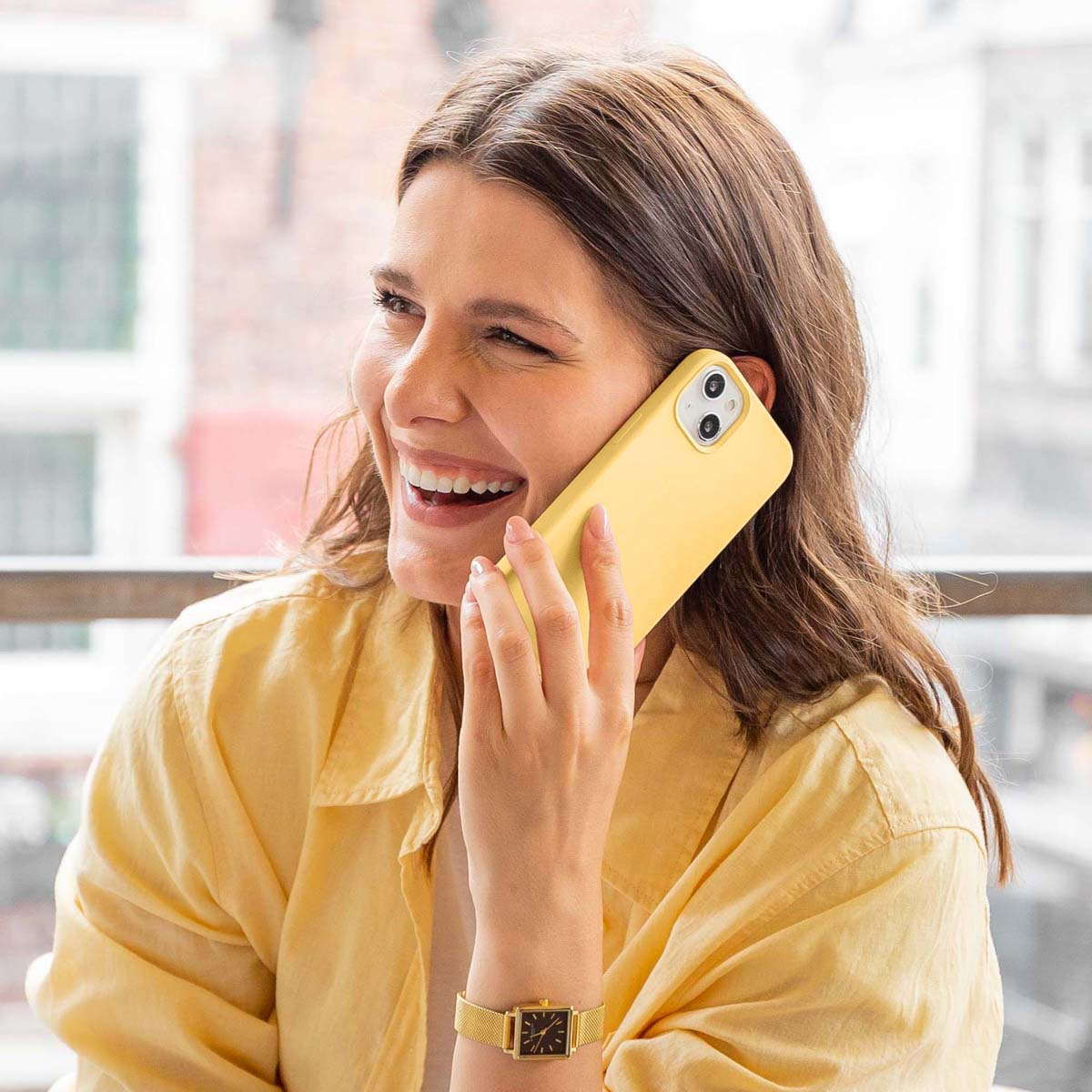 Vrouw lacht, houdt gele telefoon aan oor. Draagt gele blouse en gouden horloge.