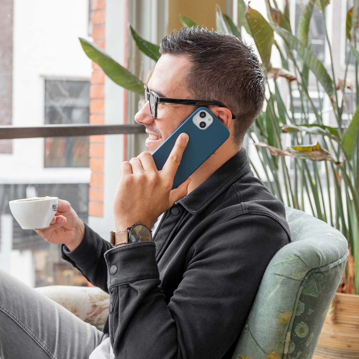 Man is aan het bellen. Hij draagt een bril en een horloge. Een kopje staat op tafel.