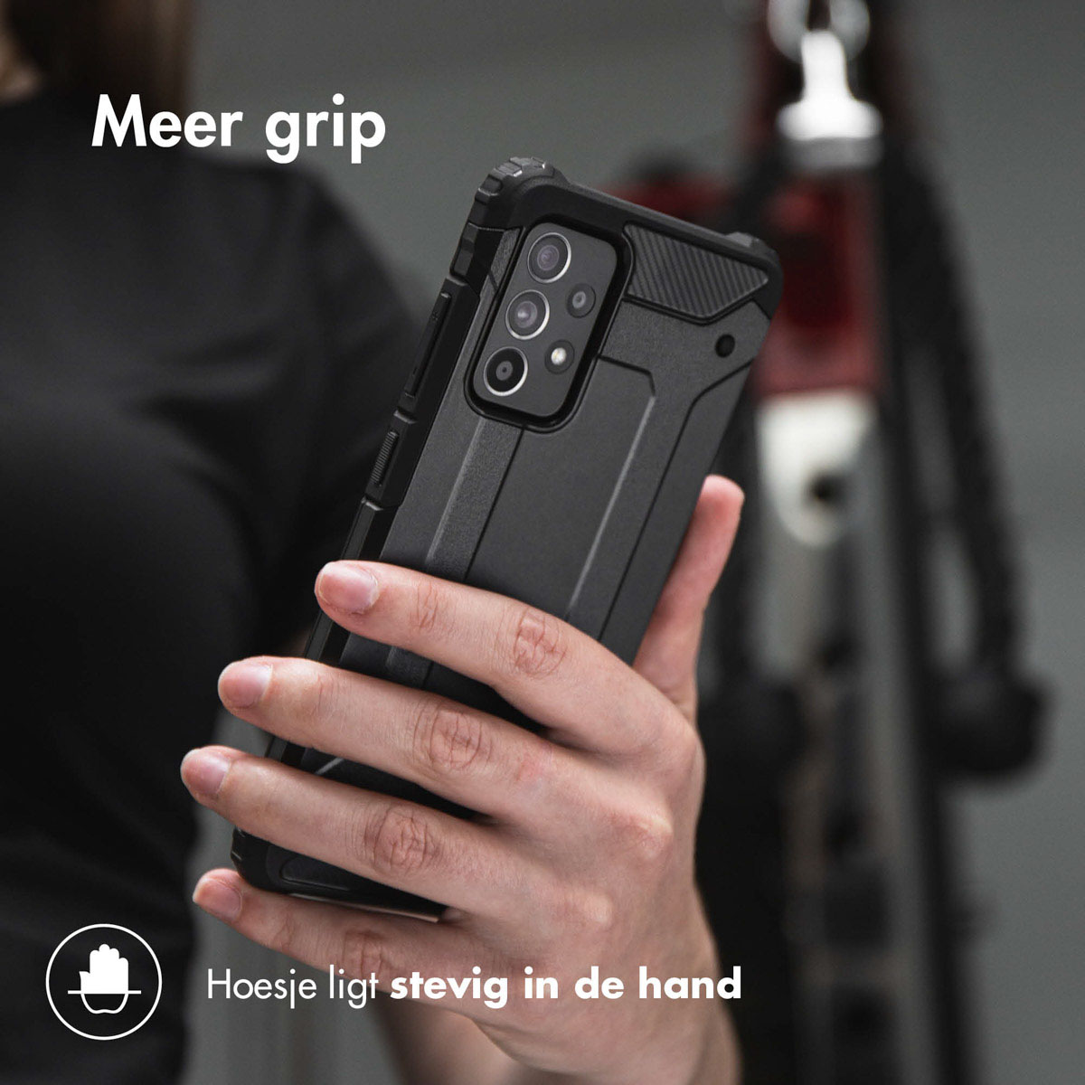 De hand van een persoon die een zwarte telefoon met een beschermhoes vasthoudt. De achtergrond toont een fiets.
