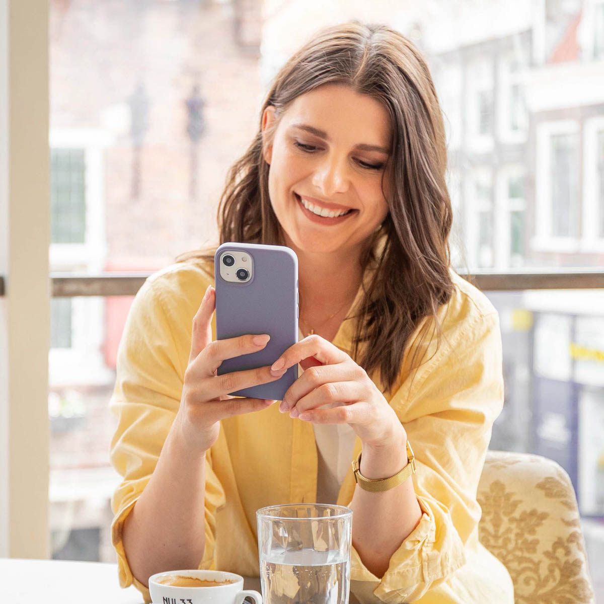 Une femme en chemise jaune sourit en regardant un téléphone violet. Il y a une tasse et de l'eau.