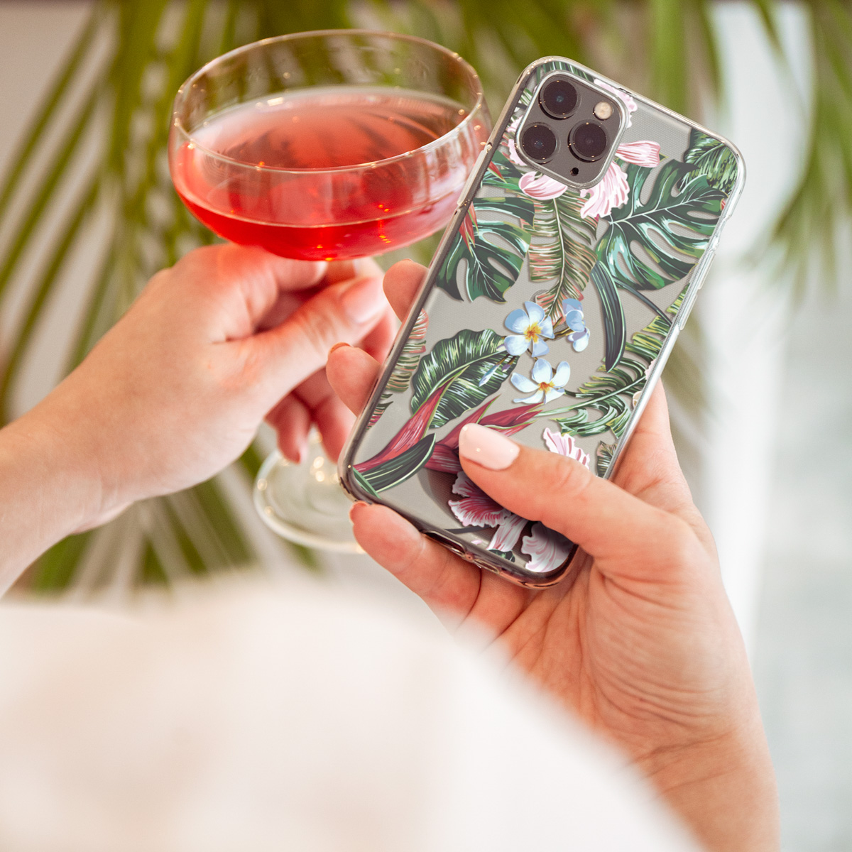 Vrouw houdt telefoon in bloemenhoes vast, met glas roze drankje en bladeren op de achtergrond.