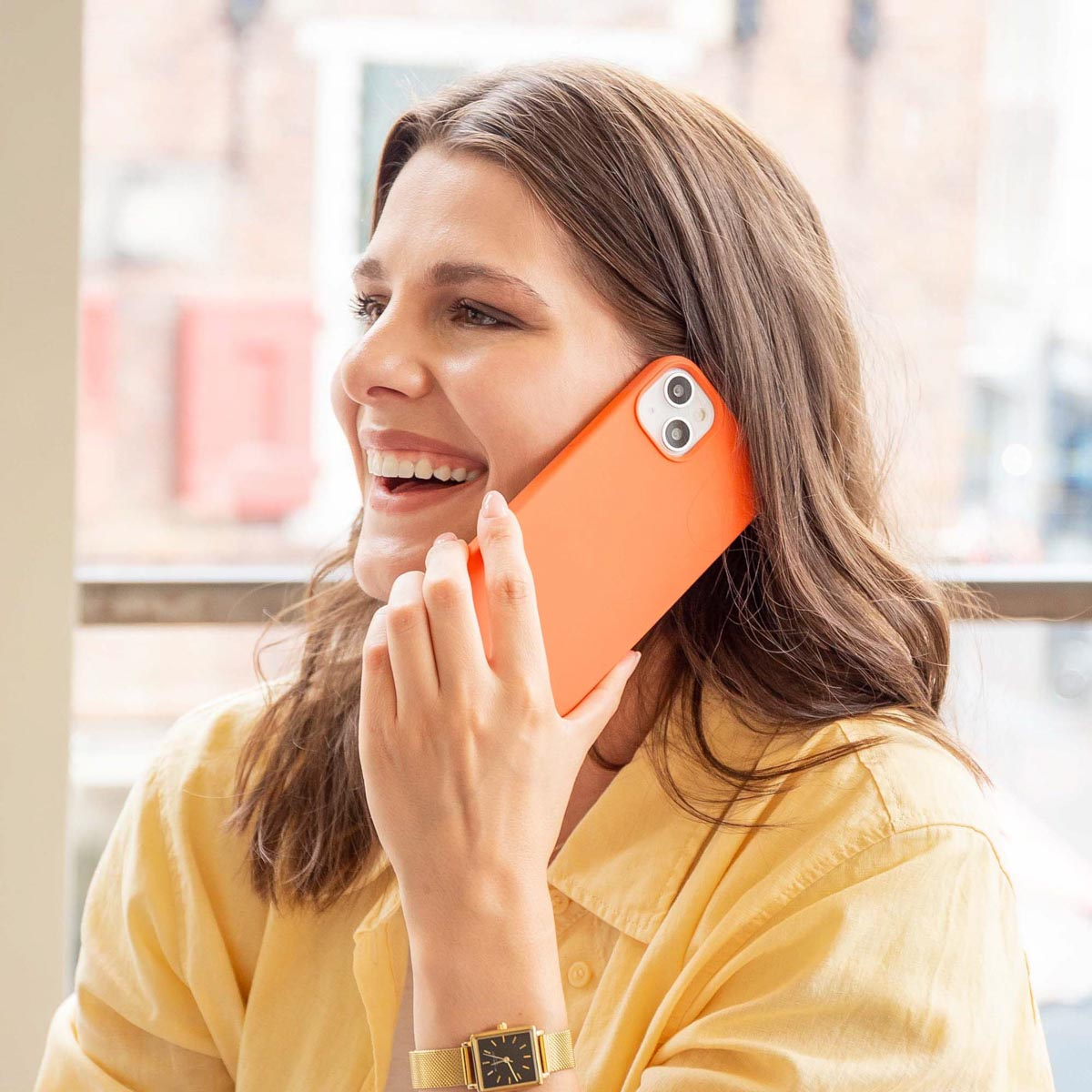 Frau lächelt, hält ein orangefarbenes Telefon ans Ohr. Sie ist draußen, mit Uhr.