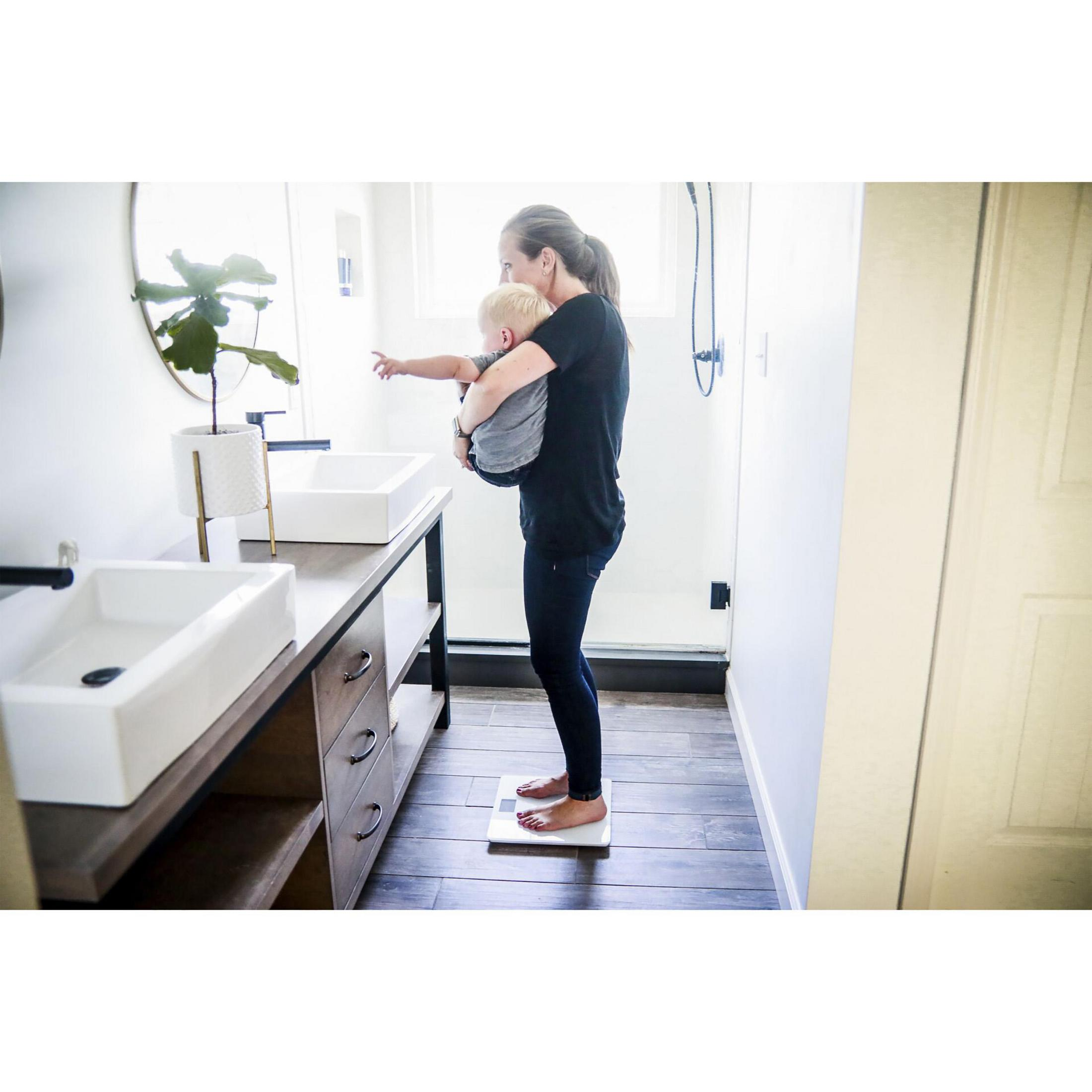 Kobieta trzymająca dziecko w łazience, stojąca na wadze. Są umywalki i lustro.