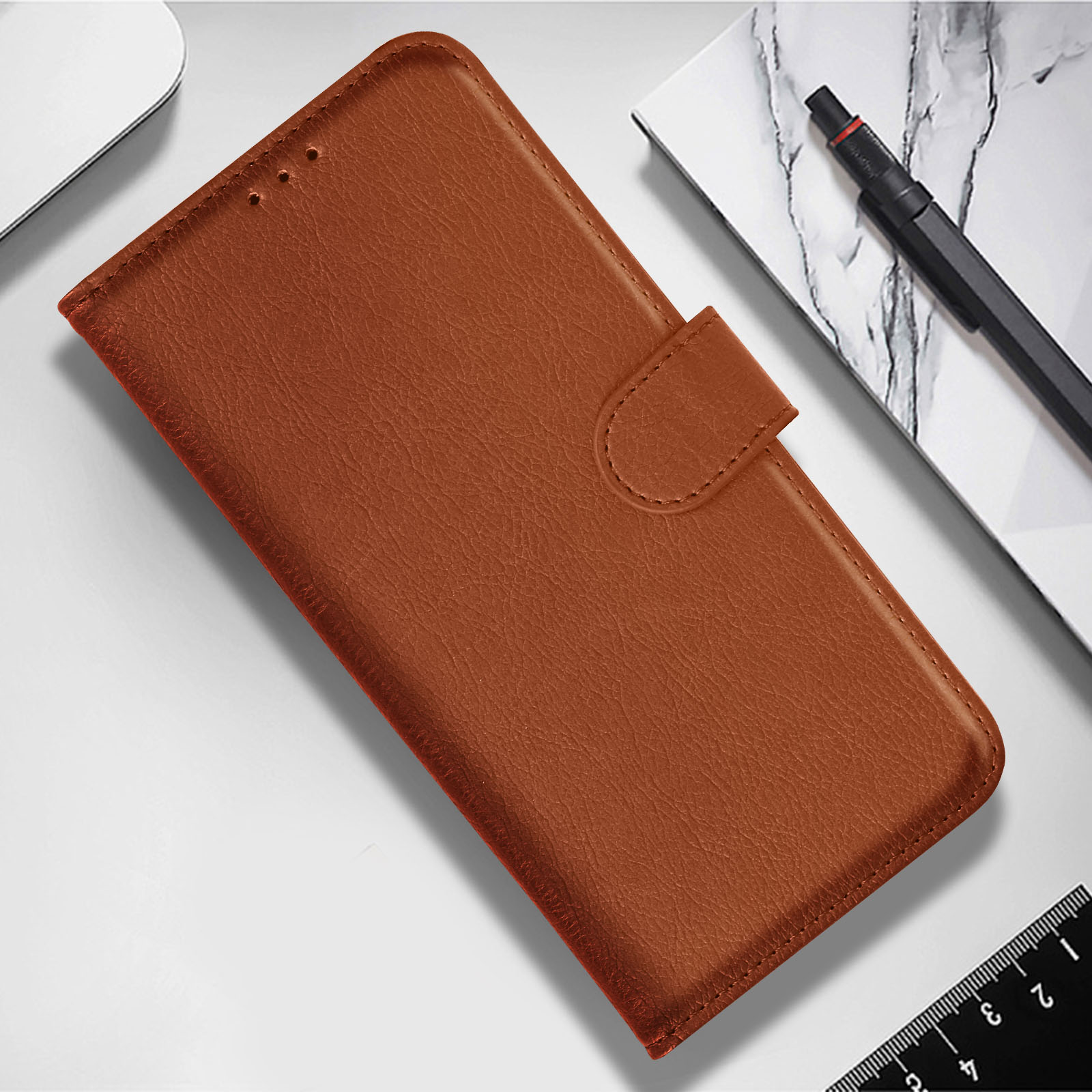 Una funda de teléfono marrón sobre una superficie blanca, junto a un bolígrafo y una regla. La funda está cerrada.