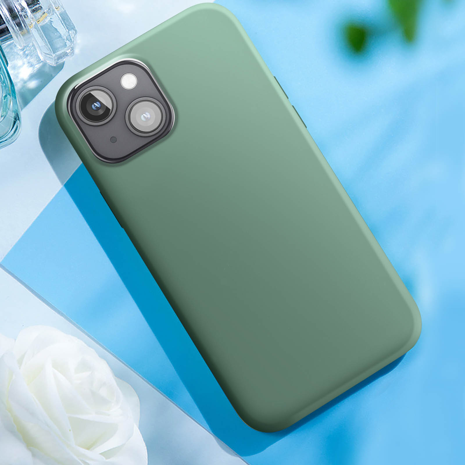 Eine grüne Handyhülle auf einer blauen Oberfläche, mit einer weißen Blume und einer Parfümflasche.