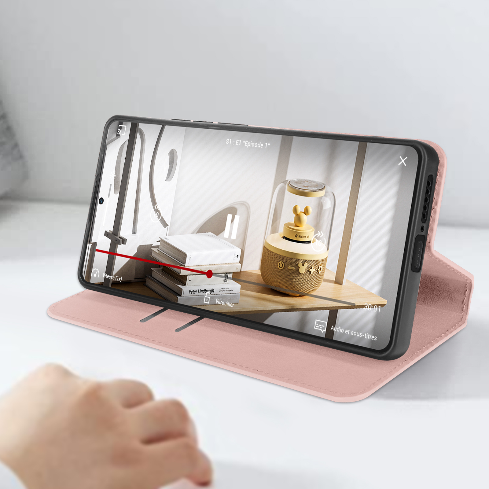 Telefono in custodia rosa, che riproduce video. Altoparlante e libri su una mensola sullo sfondo.