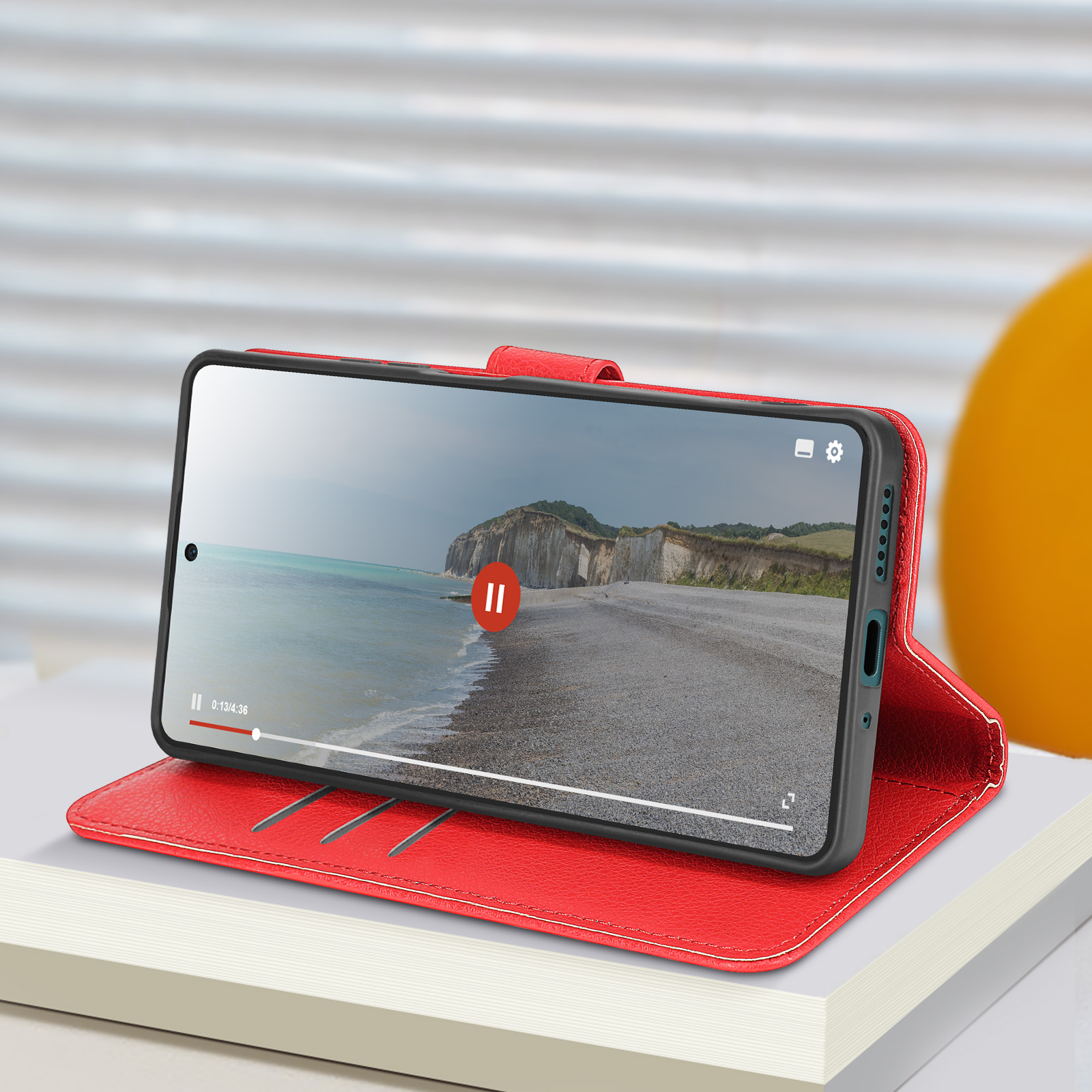 Custodia per telefono rossa con un telefono che mostra un video. La custodia è aperta, su una superficie bianca.