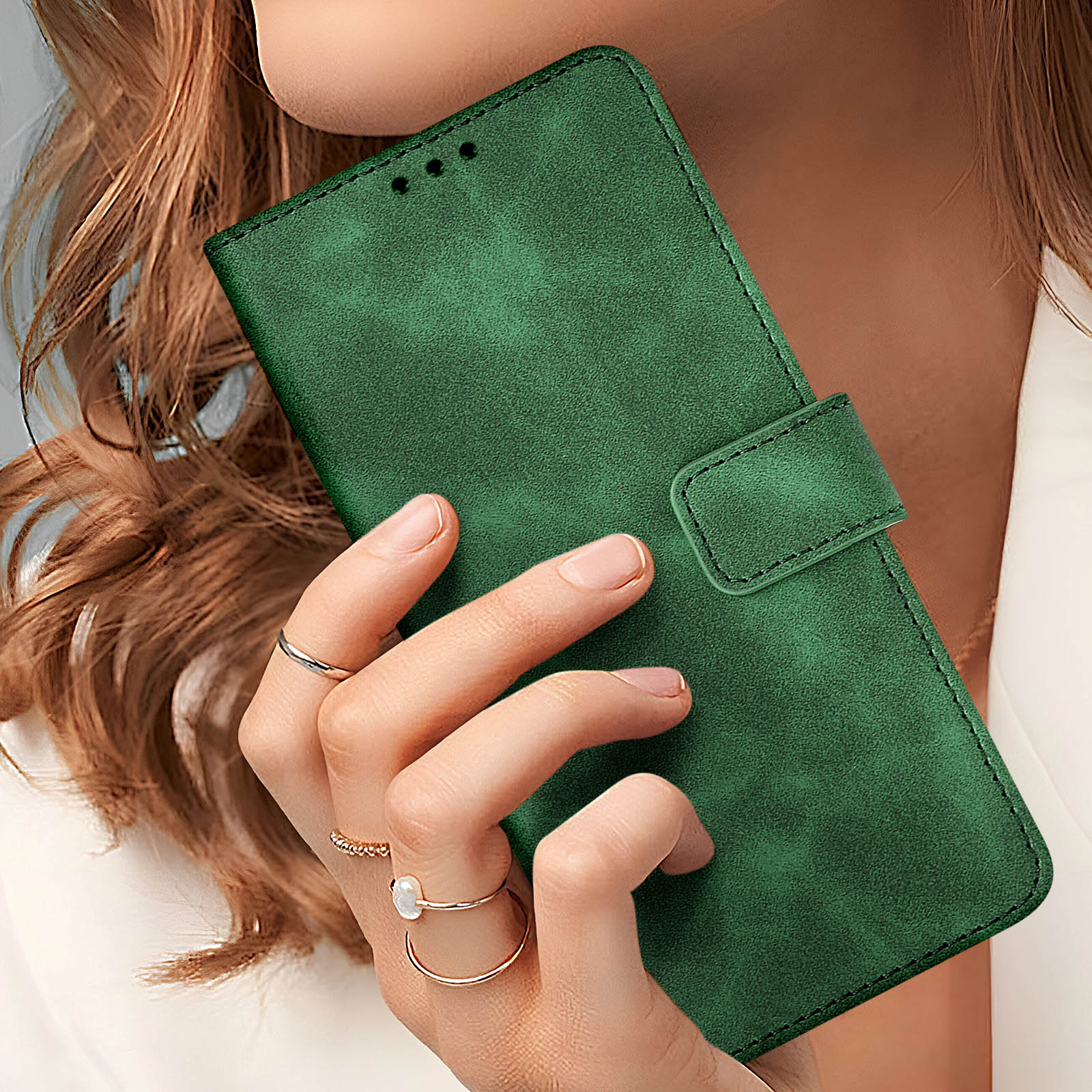Mano che tiene una custodia verde per telefono, con anelli alle dita. Sfondo giacca bianca.