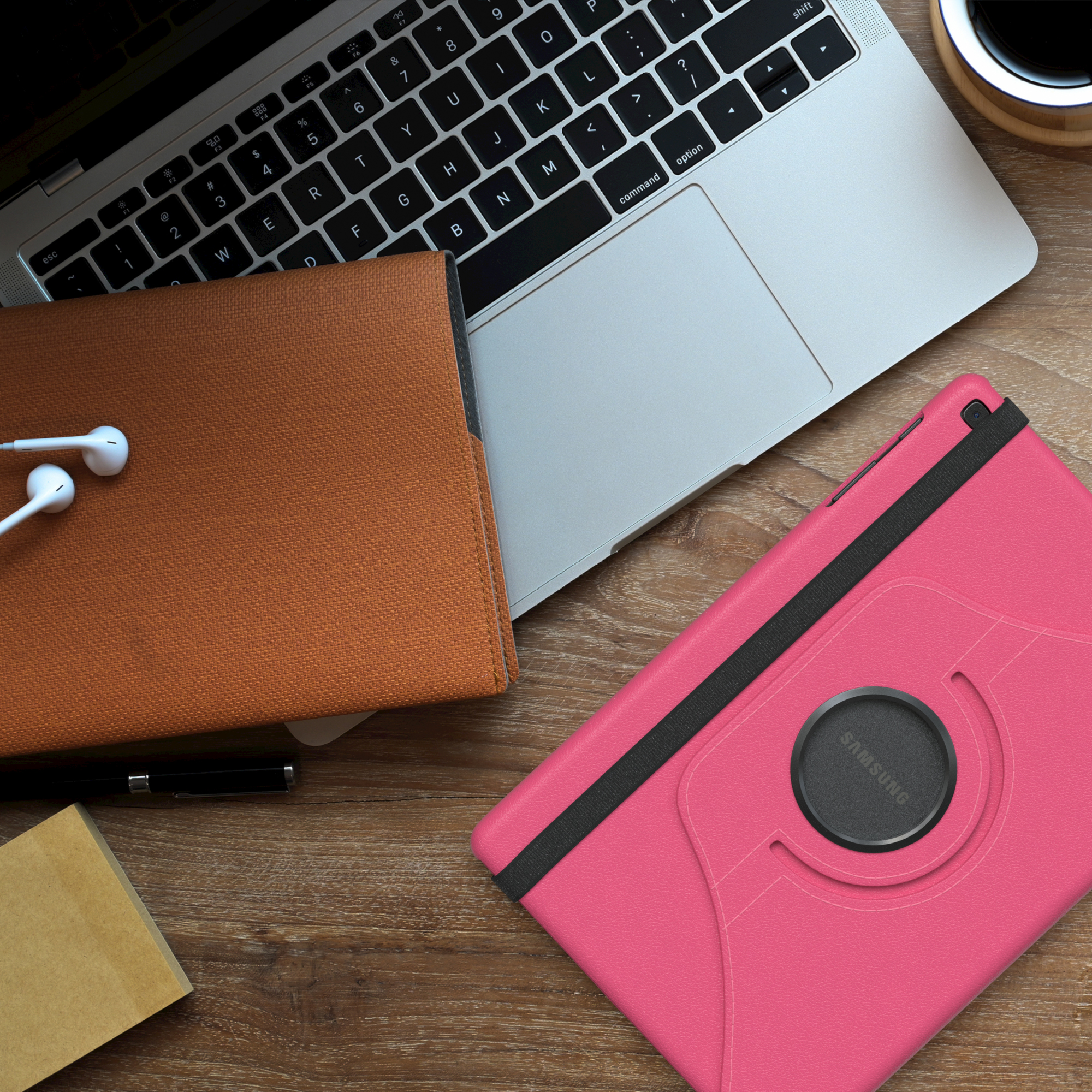Ein Laptop, Tablet in rosa Hülle, ein braunes Notizbuch und Ohrhörer auf einem Holztisch.