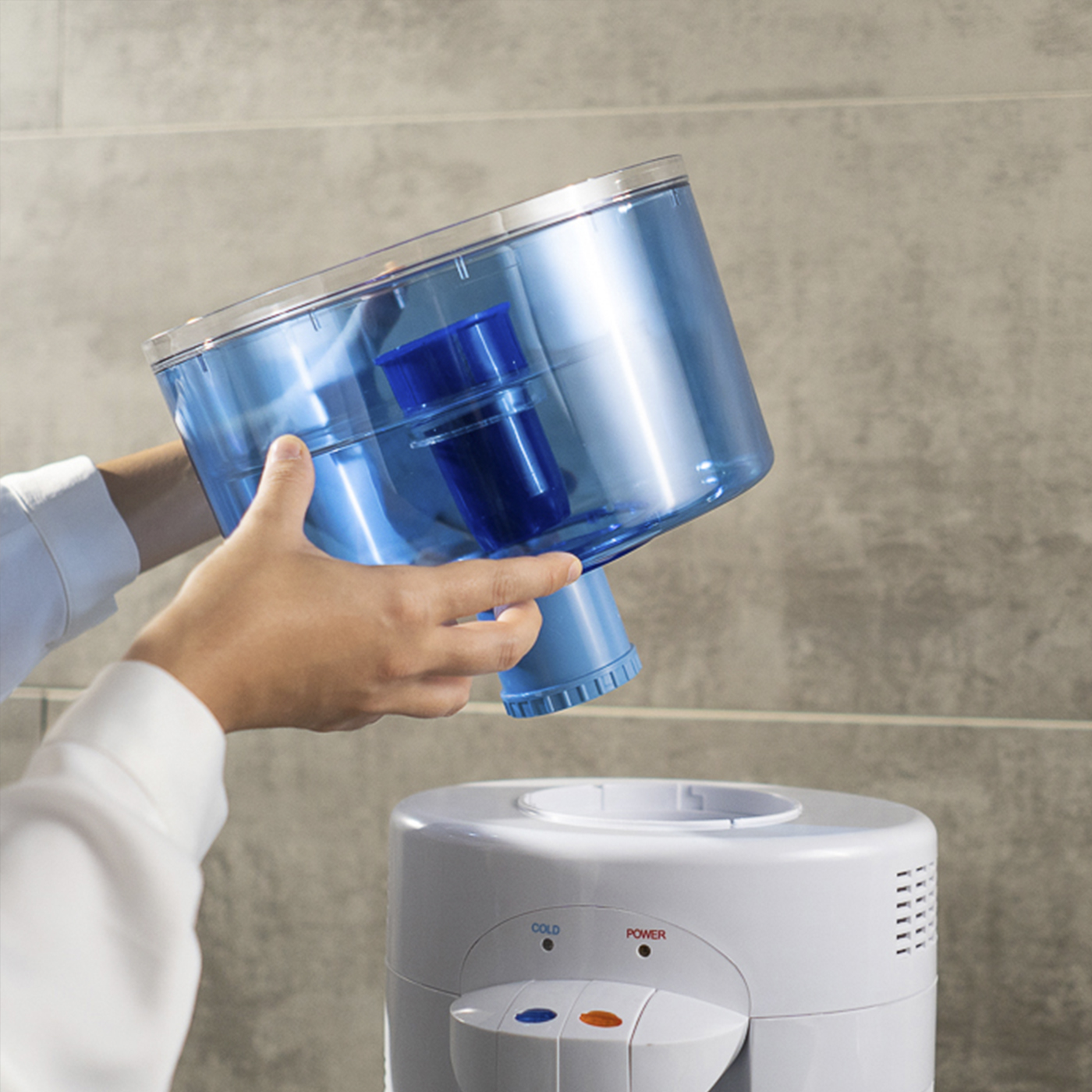 Una persona sostiene un recipiente de agua transparente y azul sobre un dispensador de agua blanco. El recipiente tiene un filtro azul.