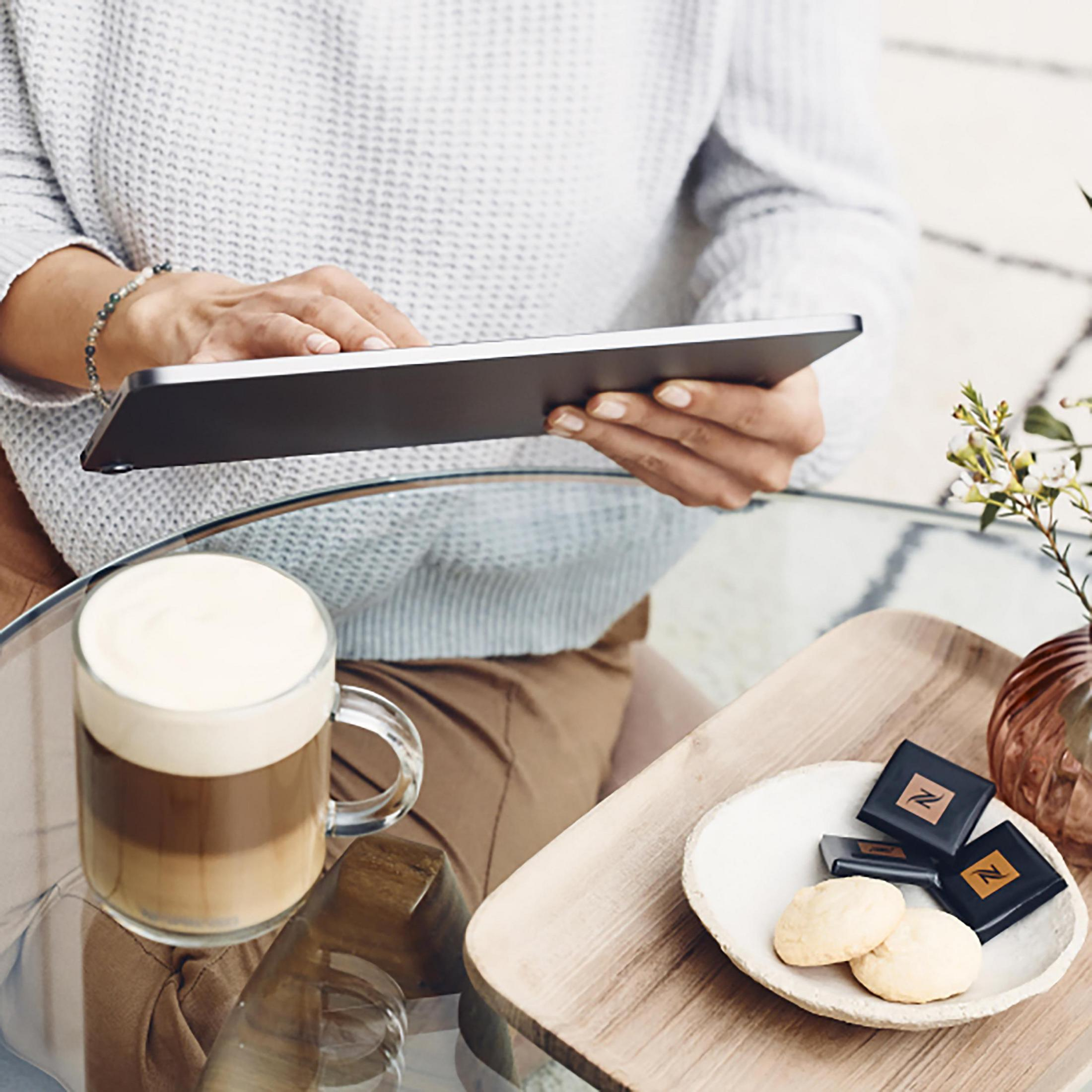 Person hält ein Tablet. Kaffee und Leckereien auf dem Tisch. Vase und Dekorationen sind in der Nähe.