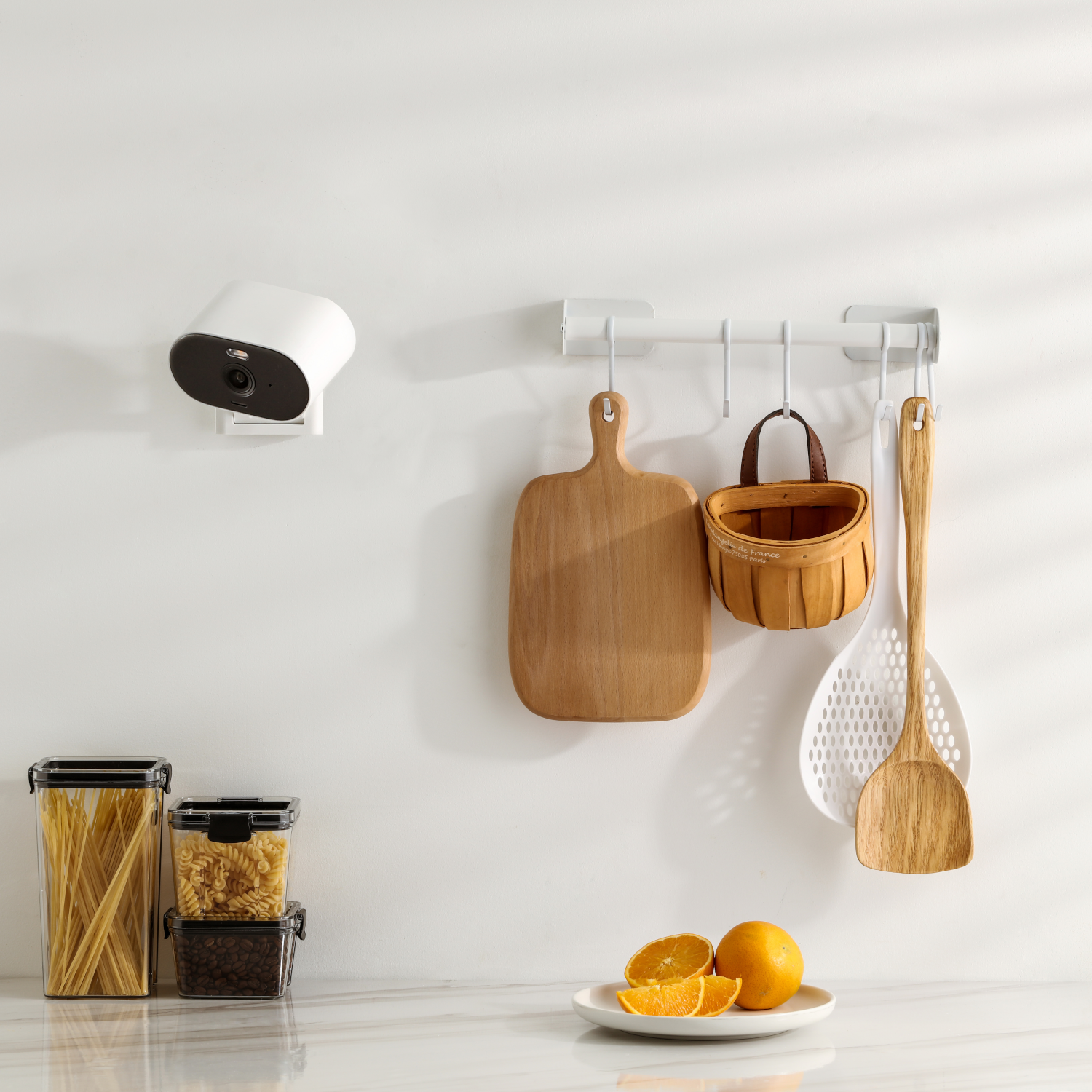 Keukenmuur met objecten: camera, snijplanken, mand, spatel, sinaasappels, potten.