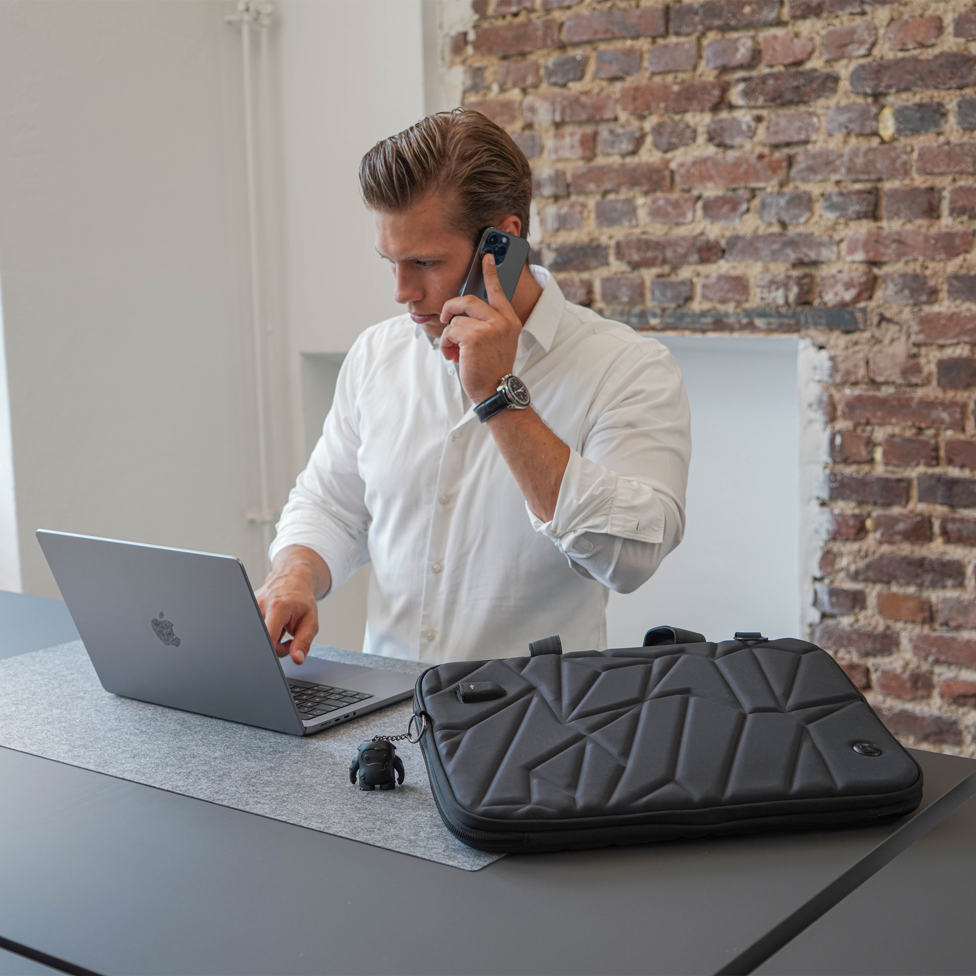 Uomo al telefono con laptop. Uomo che usa il laptop, parla al telefono. Borsa nera e muro di mattoni.