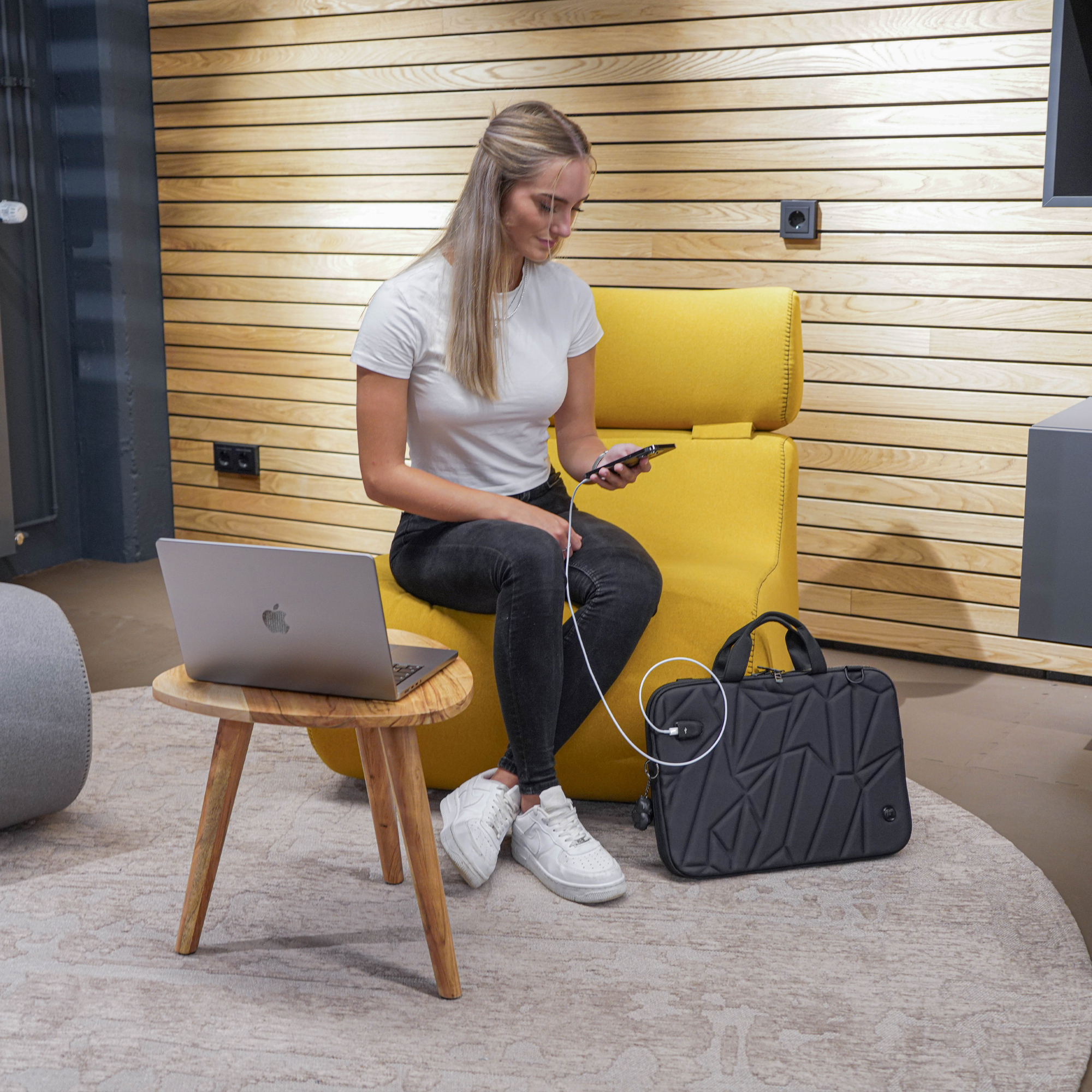 Donna usa telefono, seduta su sedia gialla, laptop su tavolo legno, borsa nera, tappeto.