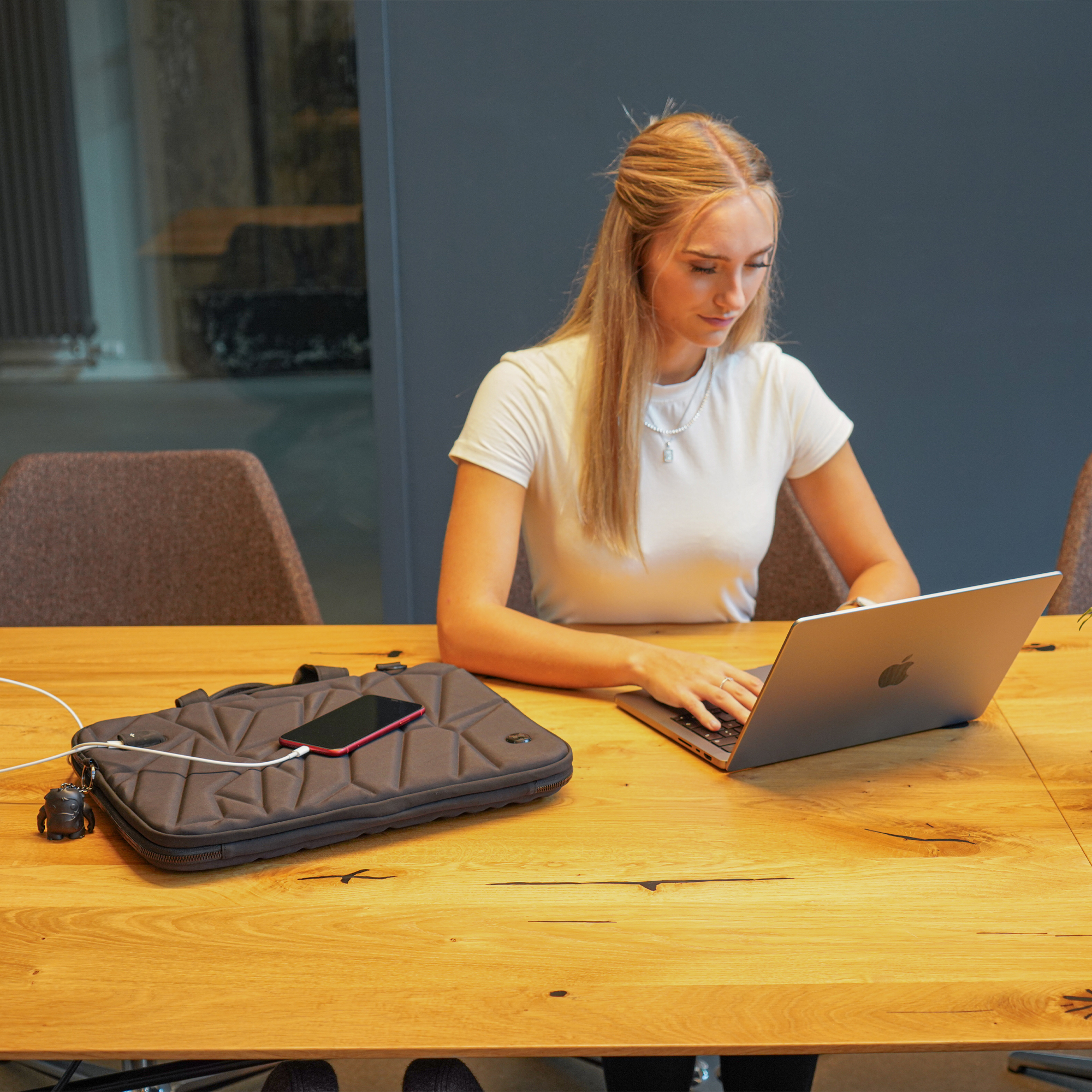 Donna che usa il laptop, telefono su custodia grigia su tavolo di legno. Ambiente ufficio.