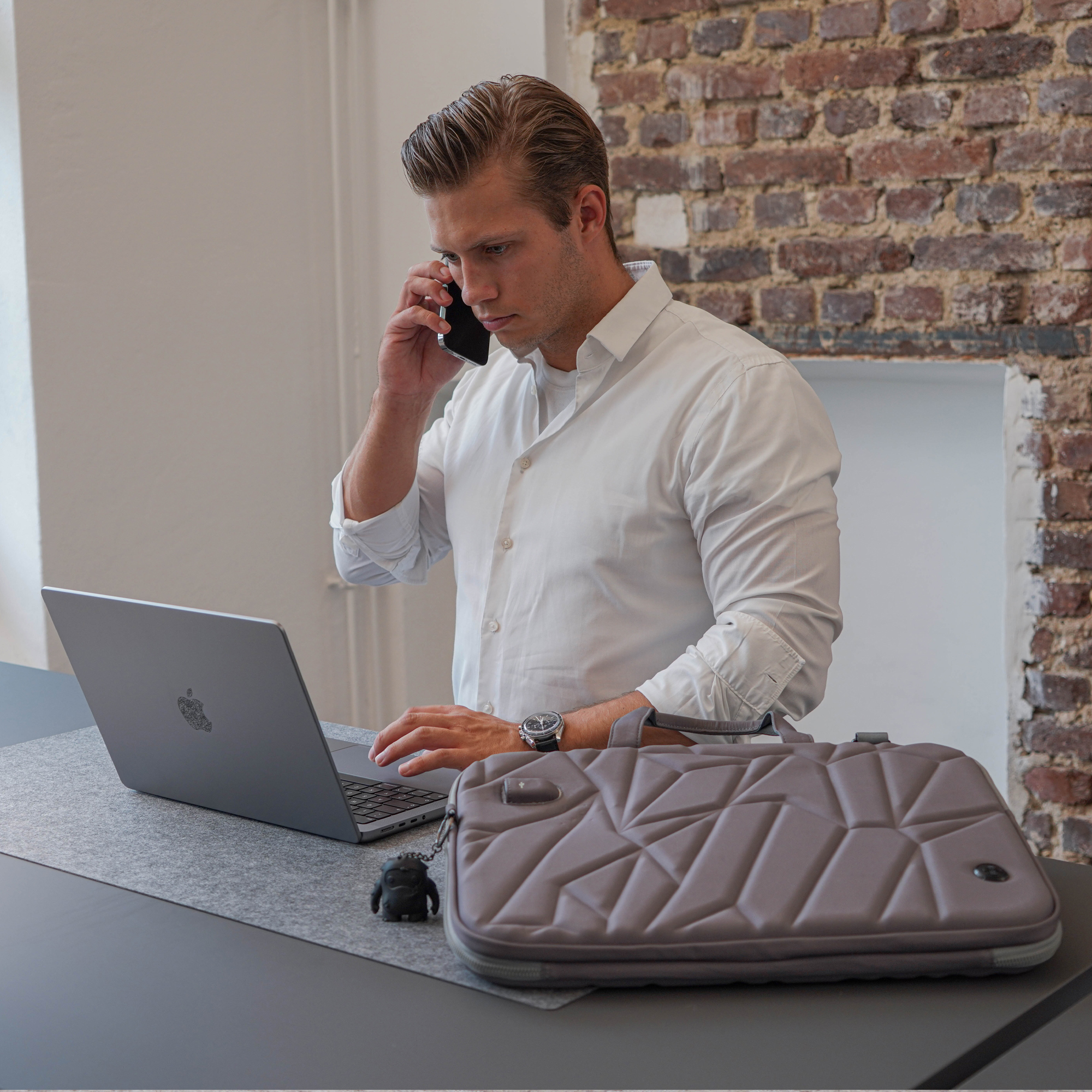 Uomo al telefono, usa un laptop. Laptop grigio, borsa marrone, figura nera.