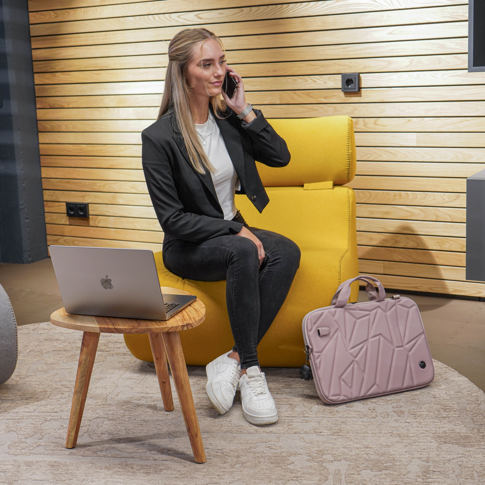 Una donna parla al telefono, seduta su una sedia gialla con un laptop e una borsa.