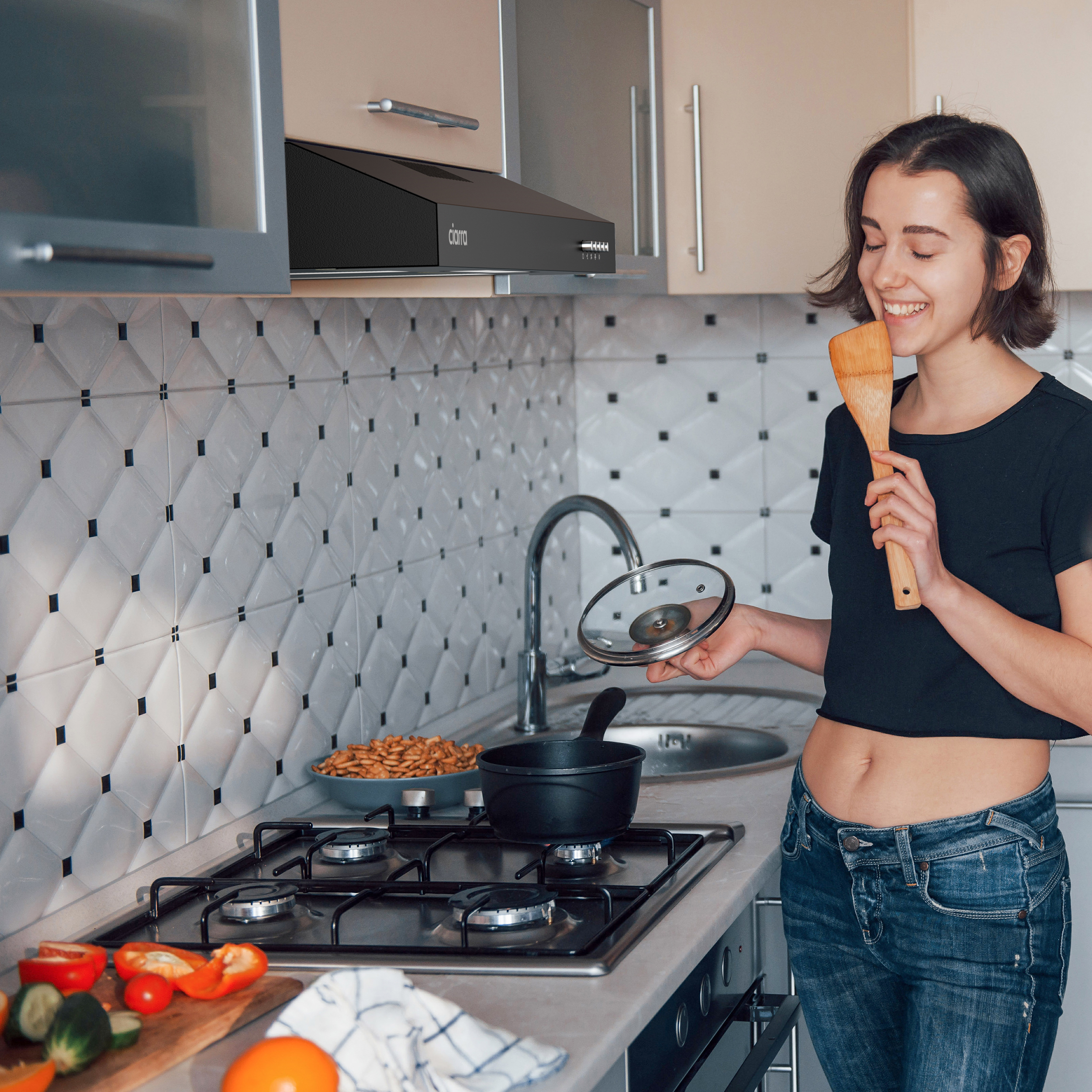 Vrouw in keuken glimlacht, houdt houten lepel en potdeksel vast, met een fornuis.