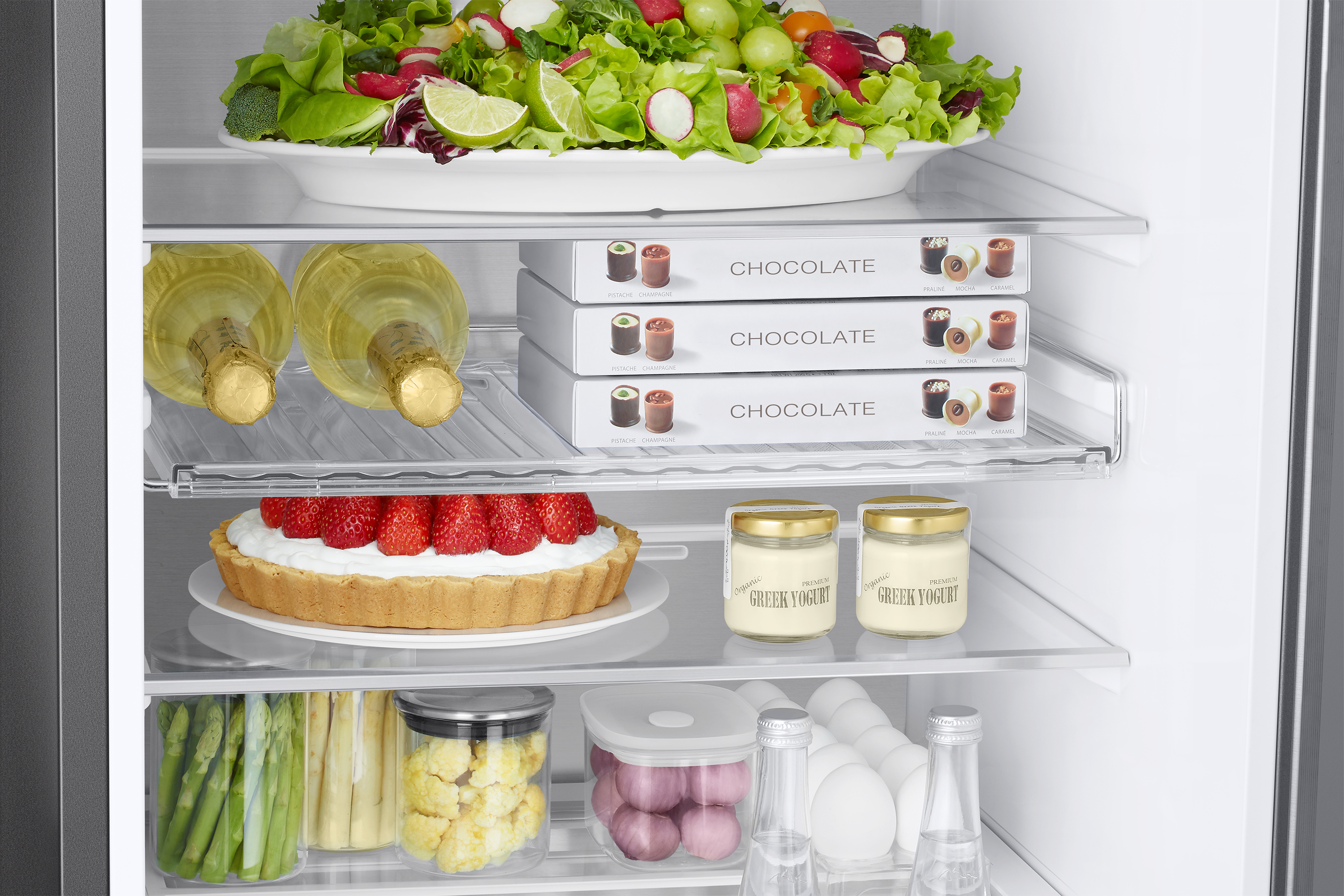 Wewnątrz lodówki z talerzem sałatki, butelkami, czekoladkami, tartą truskawkową i słoikami jogurtu.