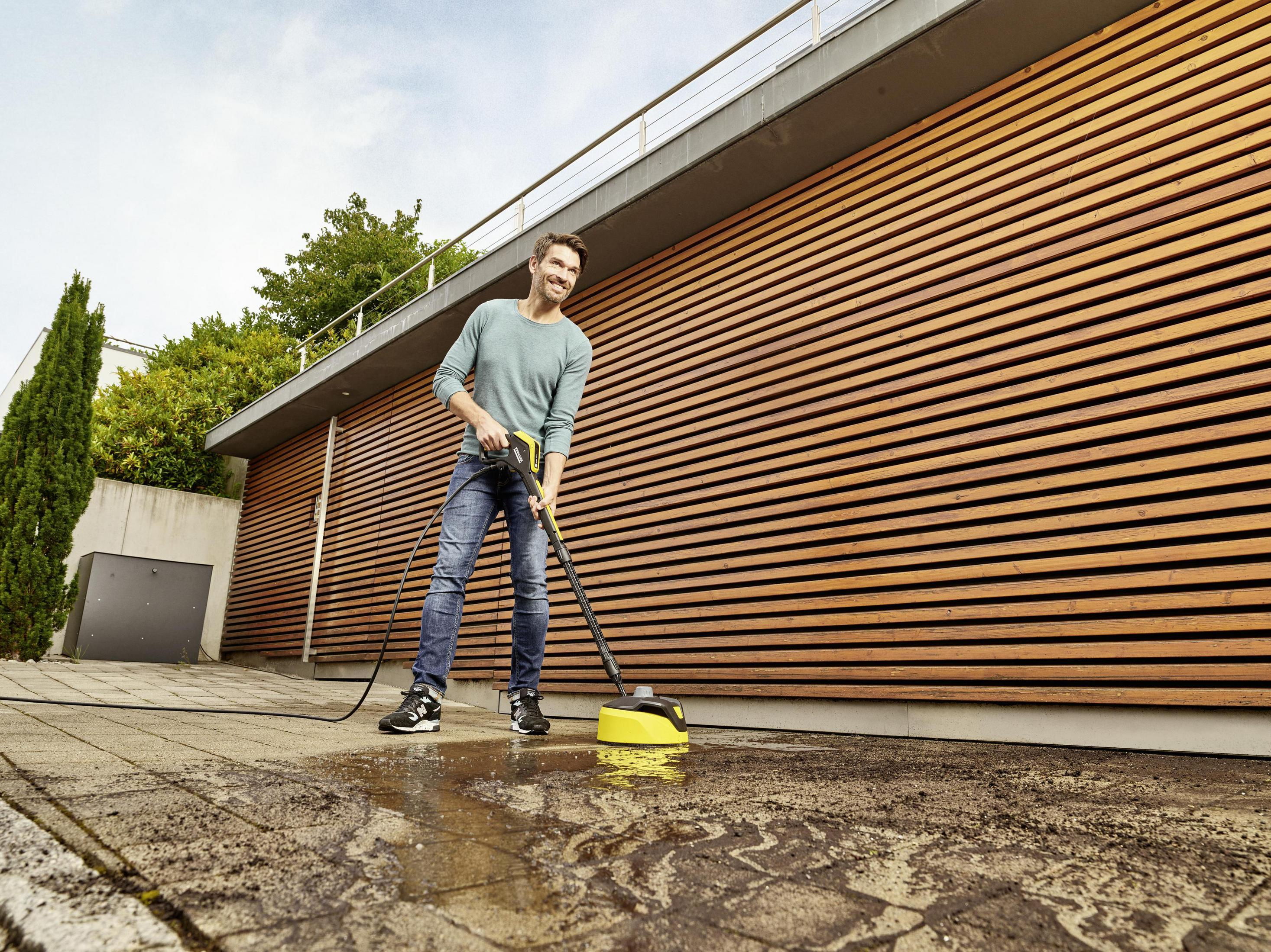 Un hombre limpiando un patio con una hidrolavadora. Pared de madera en el fondo.