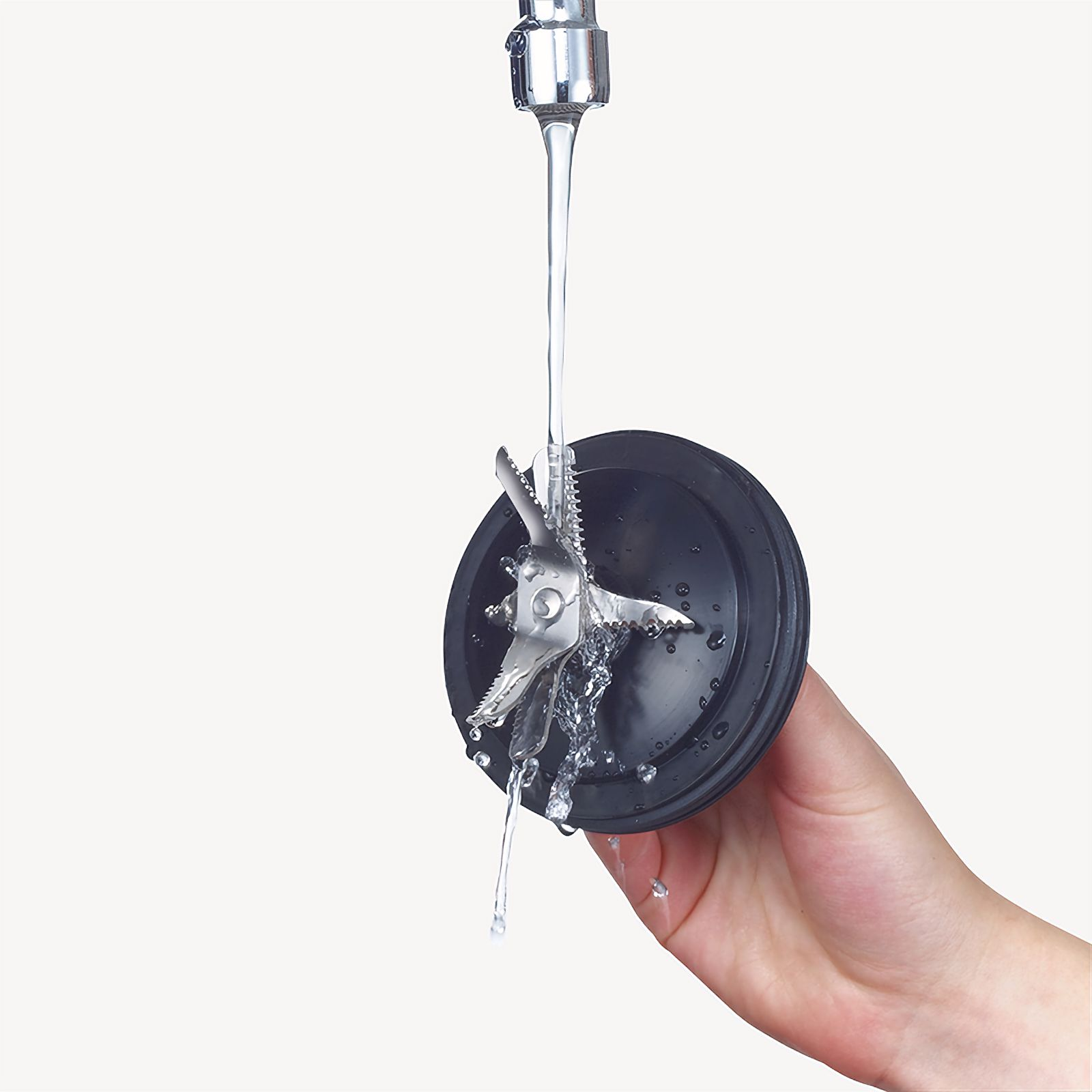 A hand holding a black blender blade under running water. Water is falling on the blades.